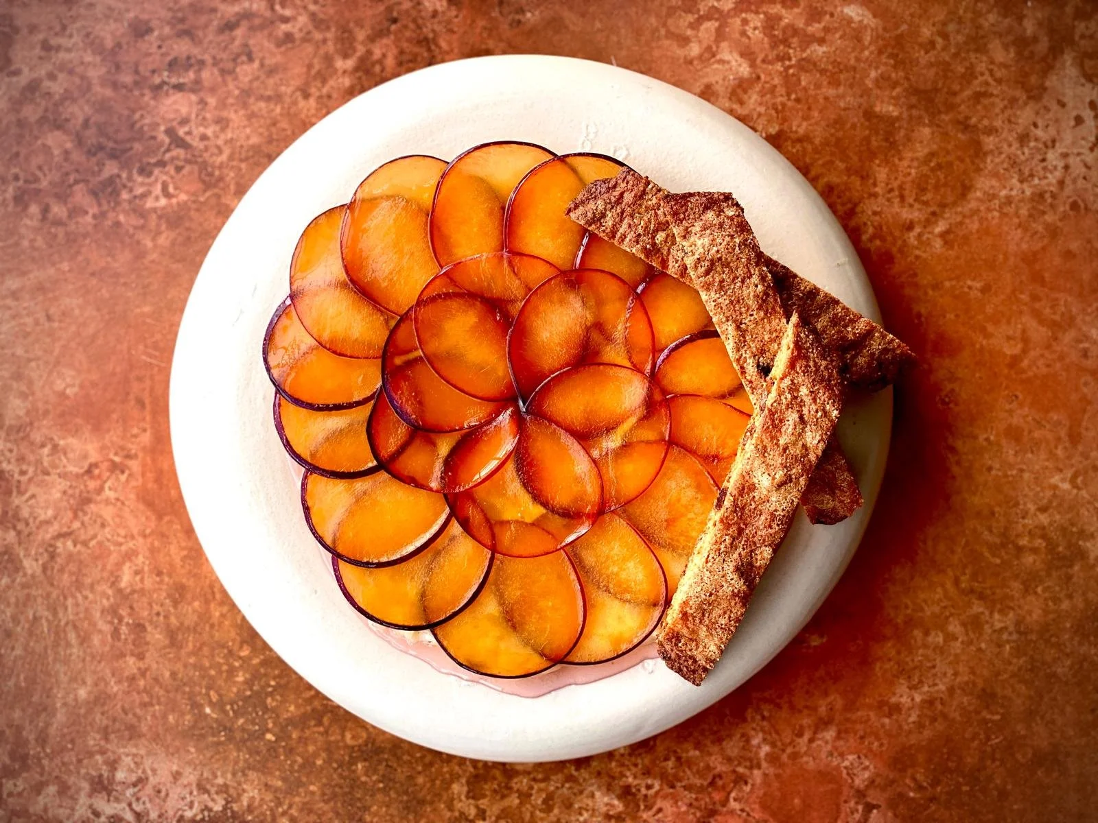 Freshly sliced orange and purple-tinted plums arranged on a white plate, garnished with a crispy pastry stick topped with powdered sugar.