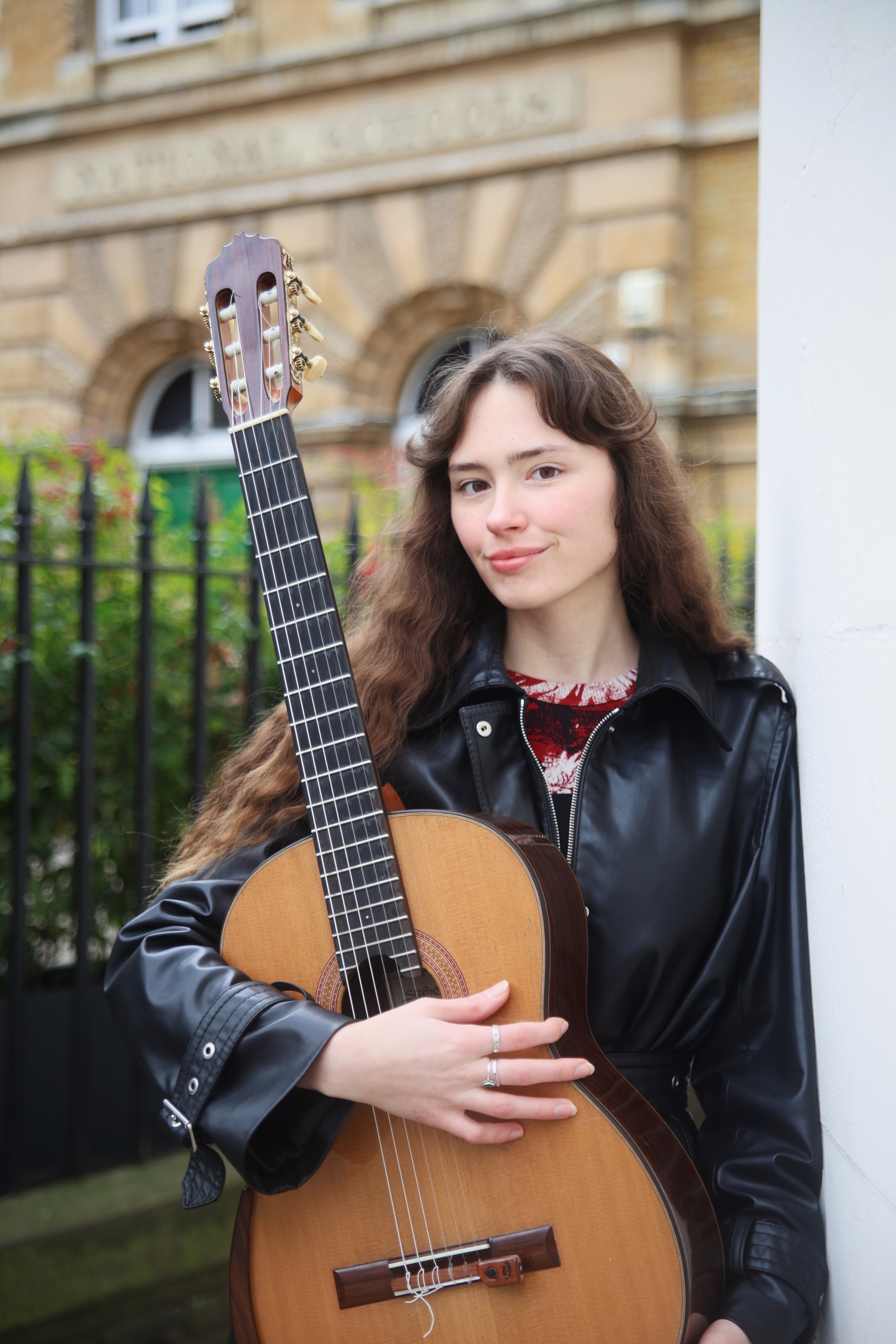 A young woman with long, wavy brown hair leaning against a white wall and holding an acoustic guitar outdoors, with a building and green bushes in the background.