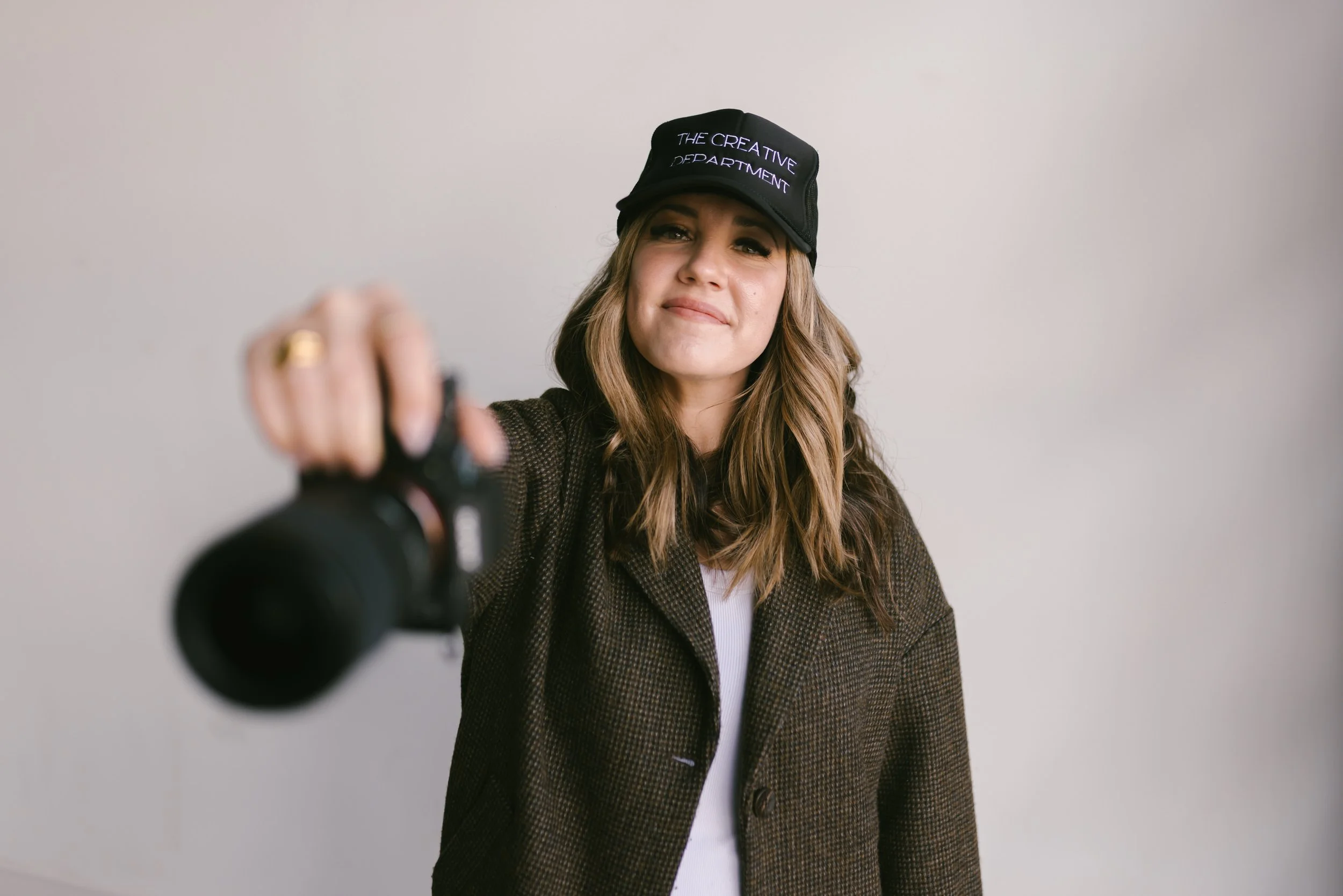 Lifestyle photographer Jules Morlet wearing a black cap that says "The Creative Department", pointing a camera towards the viewer, with a neutral plain background.