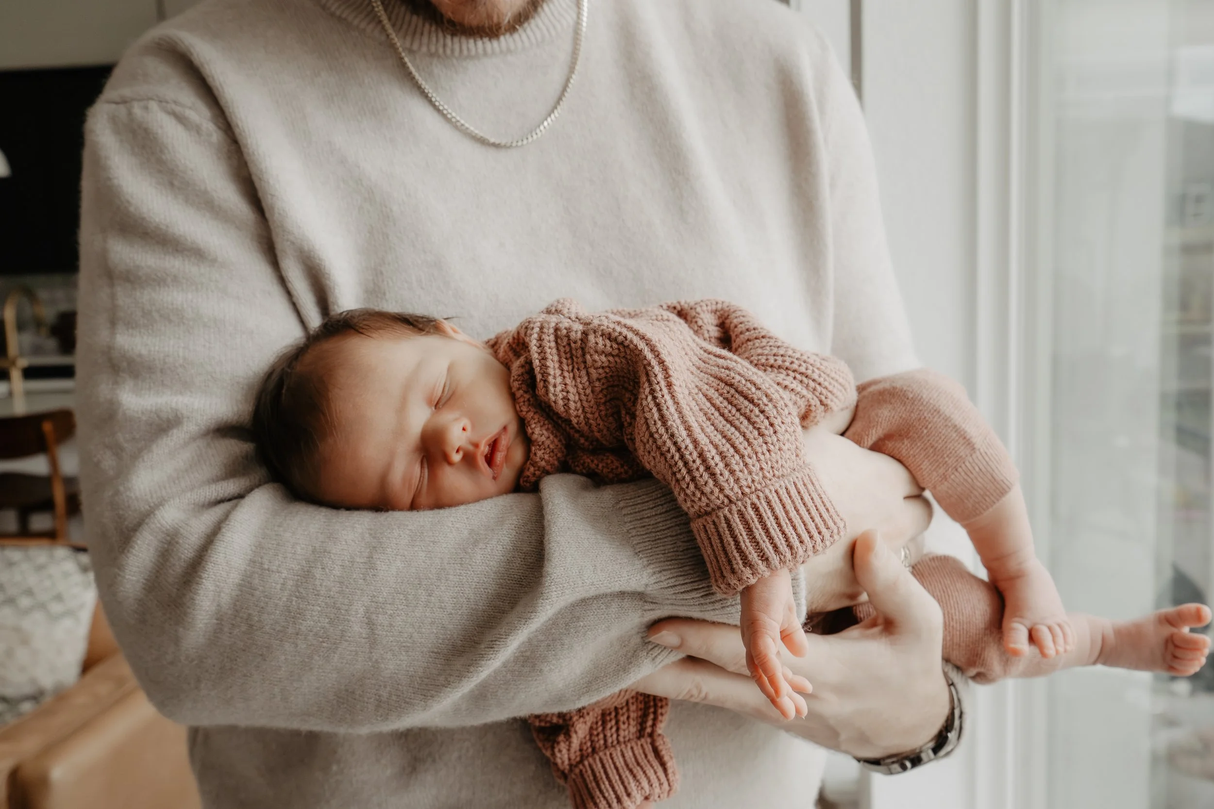 A newborn baby lies facedown, looking at the camera, asleep in their father's arms.