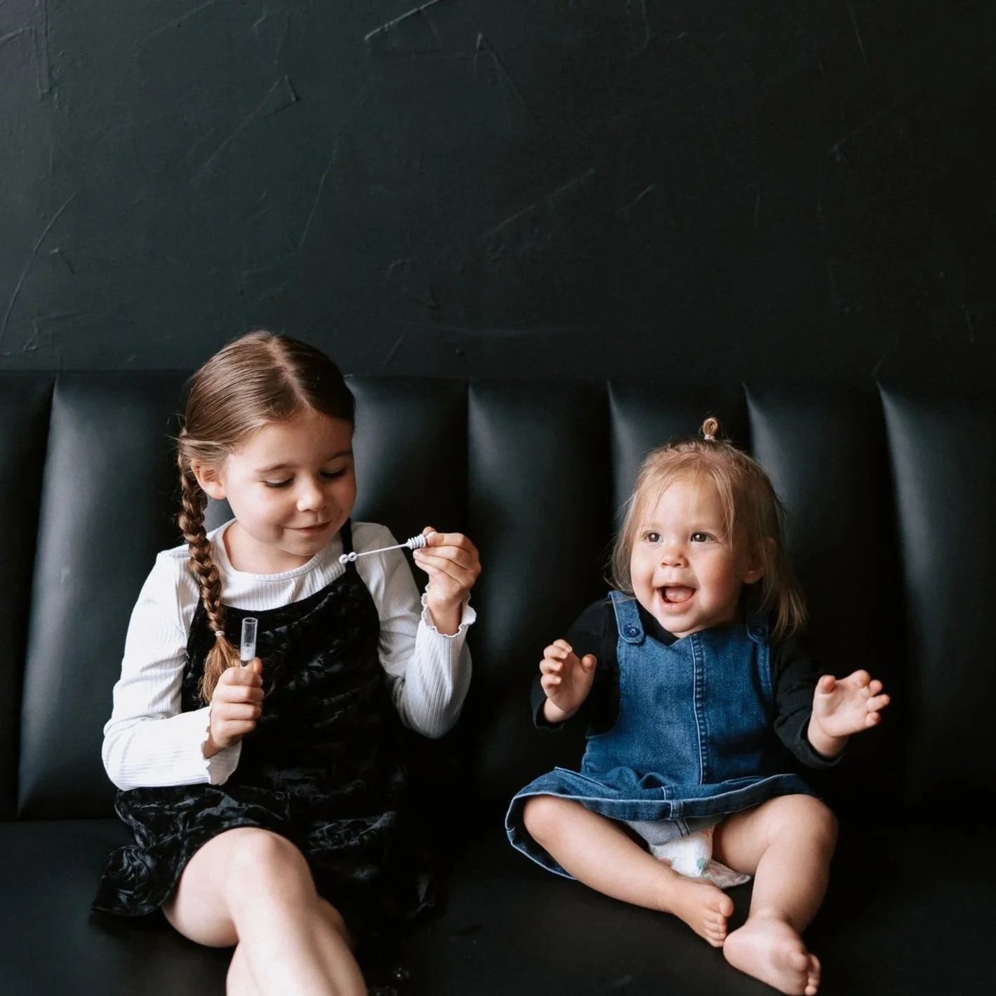 Two young girls sitting on a black bench, one with braided hair holding a bubble wand, and the other with blond hair, wearing a denim dress, smiling and clapping.