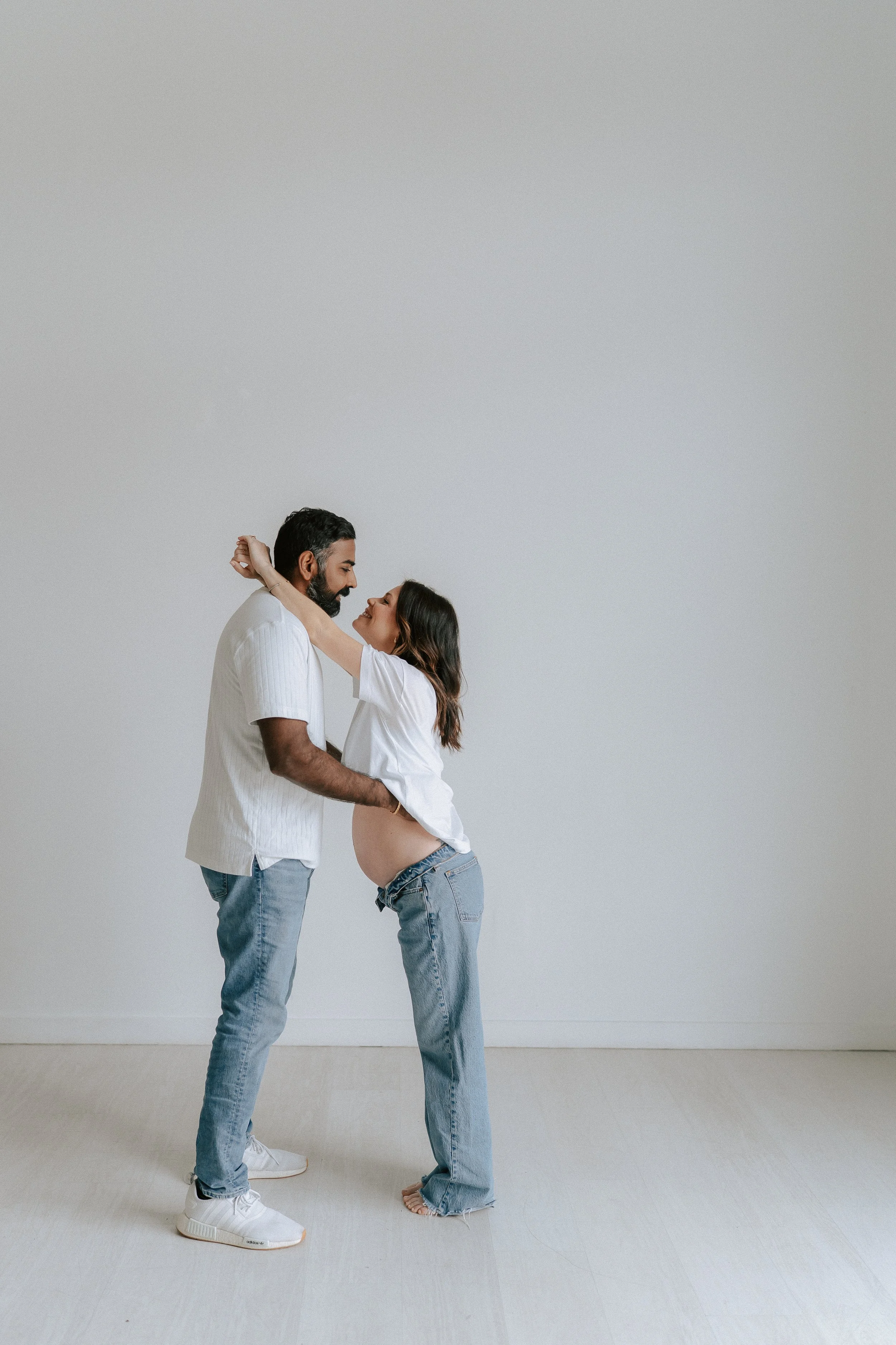 An expecting couple standing in an embrace in front of a white studio wall