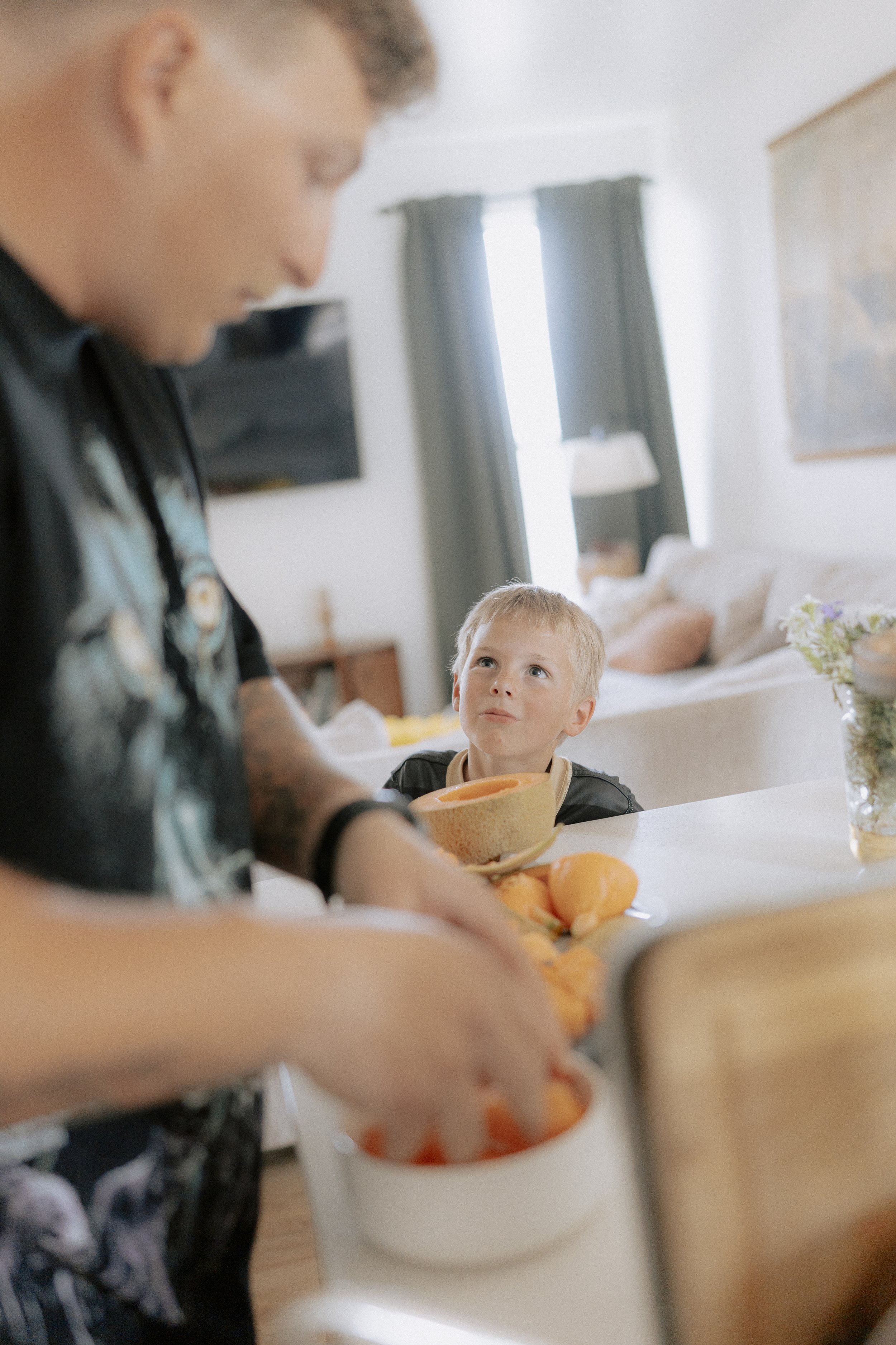 A young boy with blonde hair looking up at an adult as they prepare food with oranges on the counter in a bright room with curtains and artwork on the walls.