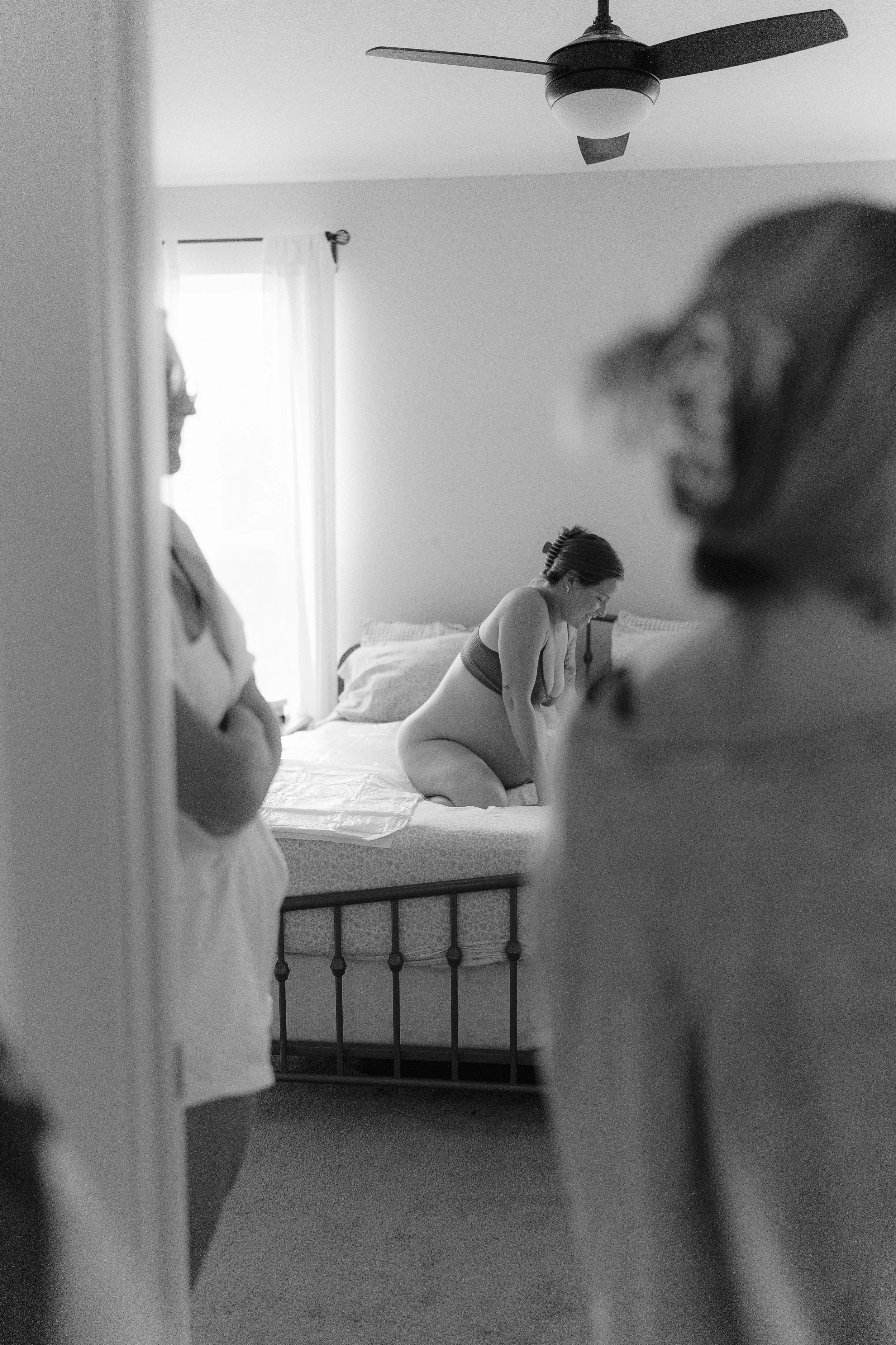 A woman sitting on a bed in a bedroom, with two other women standing nearby, viewed through a doorway in black and white.