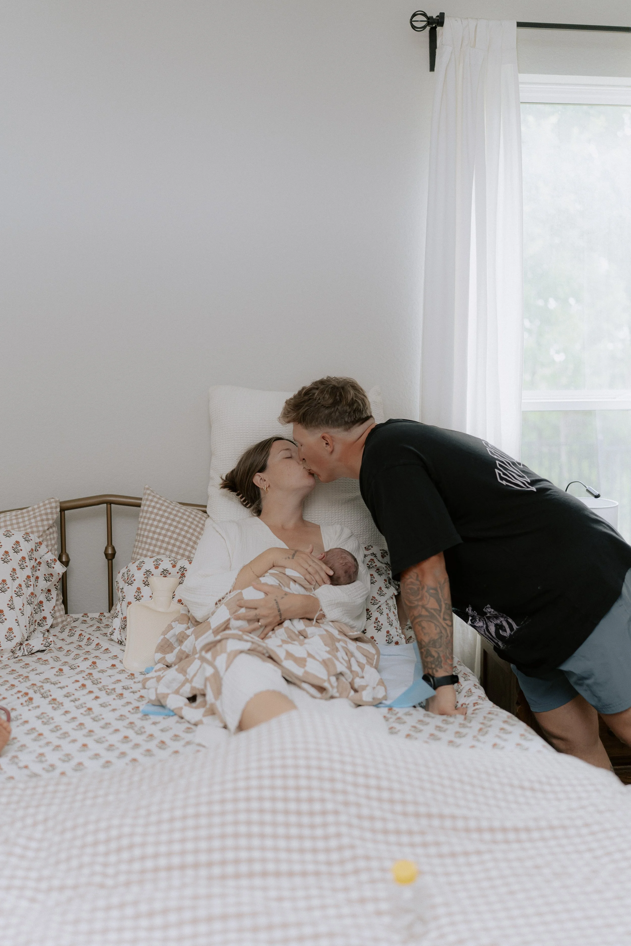 Woman holding a newborn baby in bed, while a man is leaning over and kissing her on the forehead.