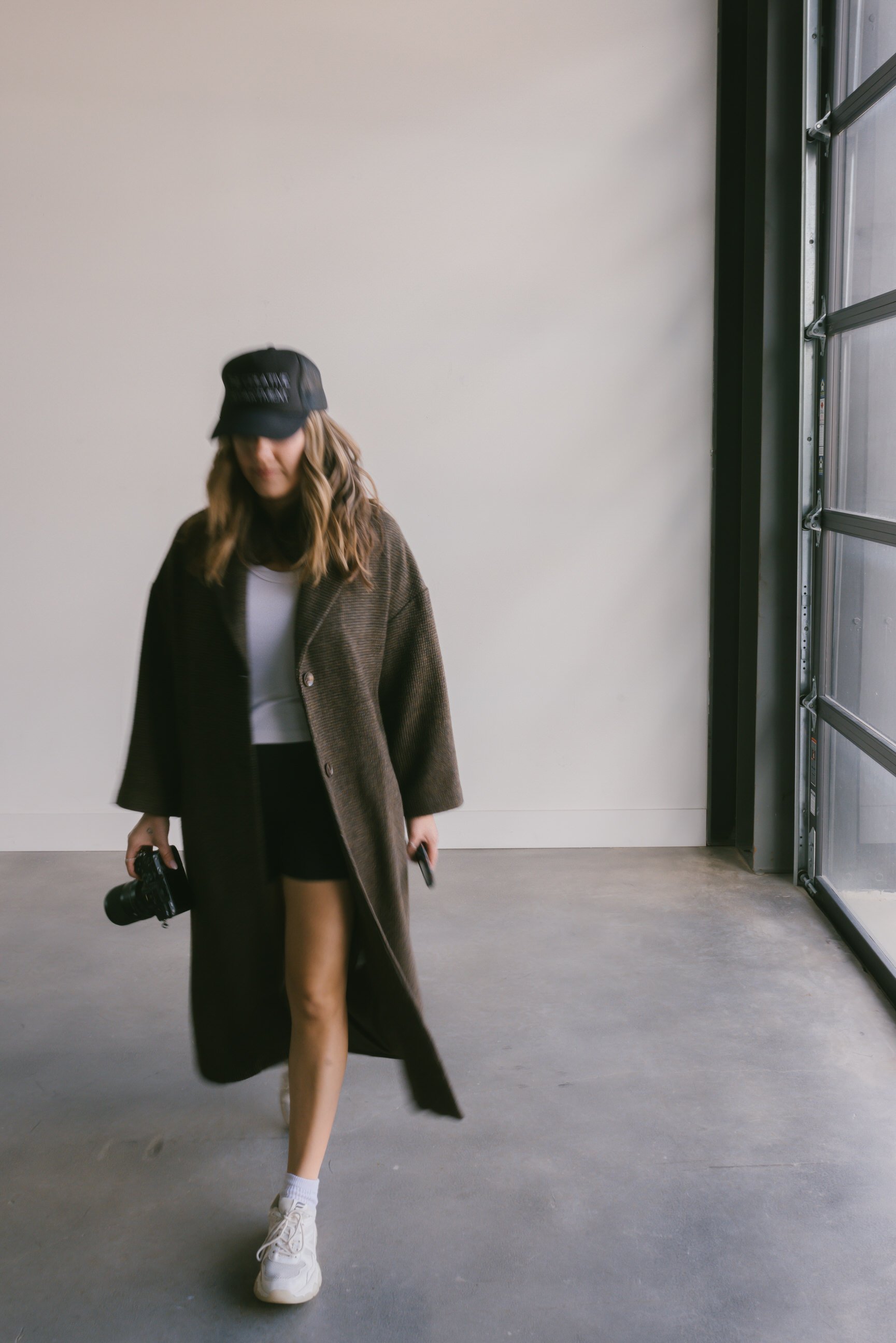 Austin Based maternity photographer Jules Morlet standing on a concrete floor, holding a camera and a phone, wearing a long brown coat, white top, black shorts, white sneakers, a black cap, and has wavy hair, inside a modern room.