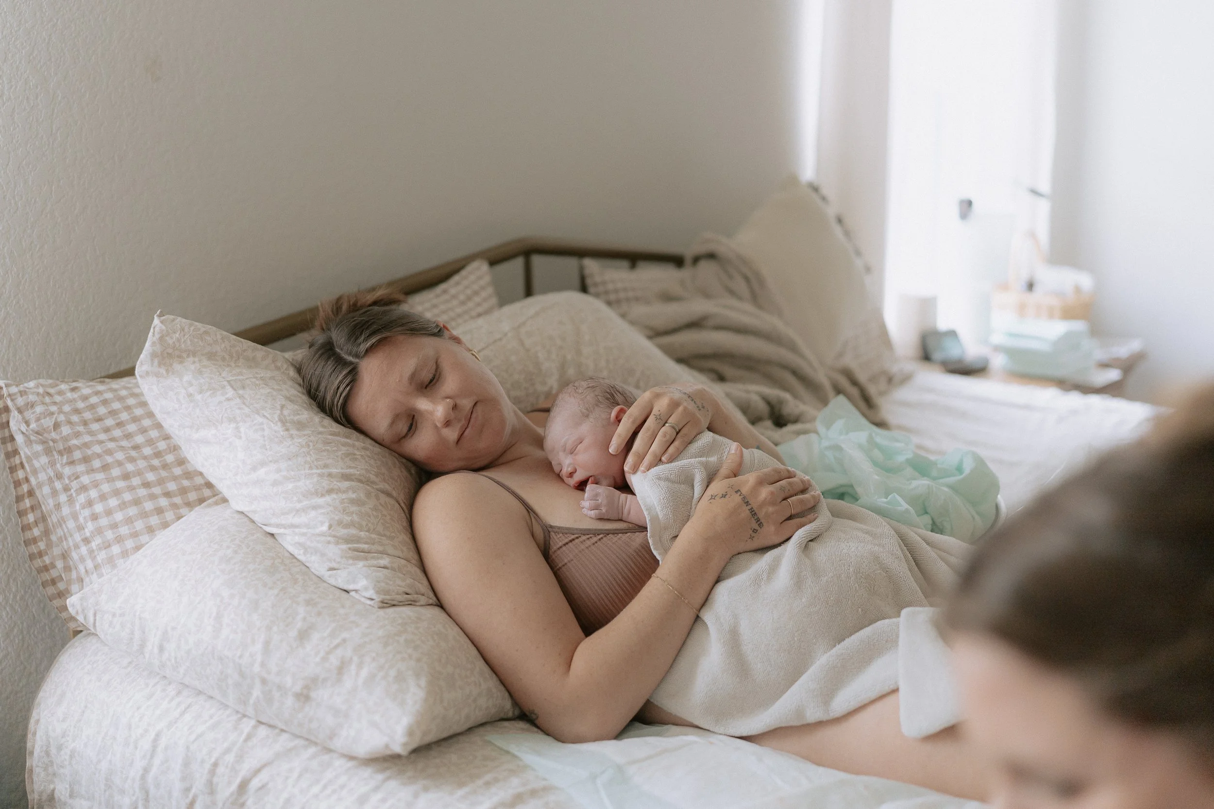 A woman with gray hair and tattoos is lying in bed holding a newborn baby close to her chest, both sleeping peacefully.