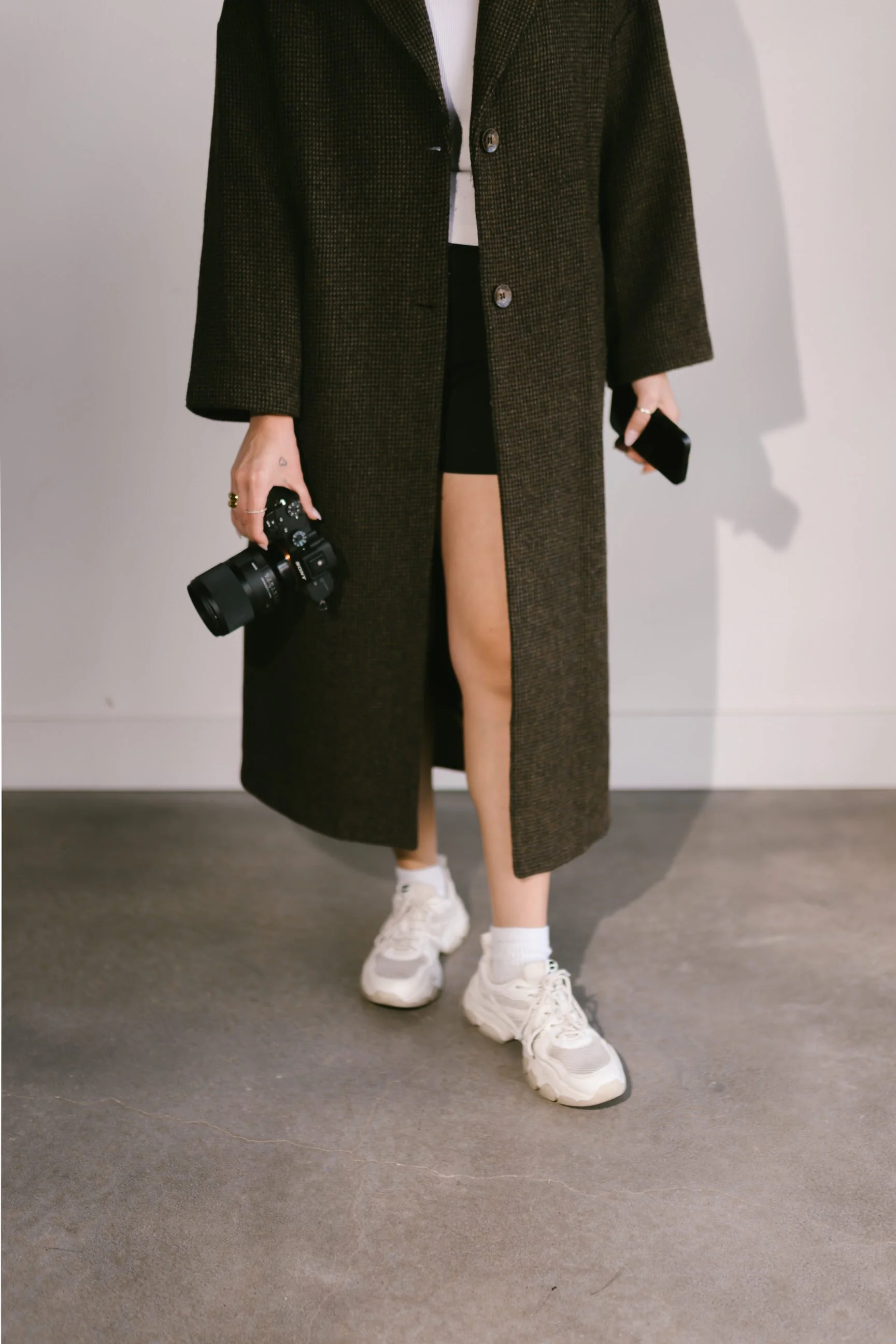 Lifestyle photographer Jules Morlet wearing a long dark coat, black shorts, white sneakers, and holding a camera and phone, with one leg exposed.