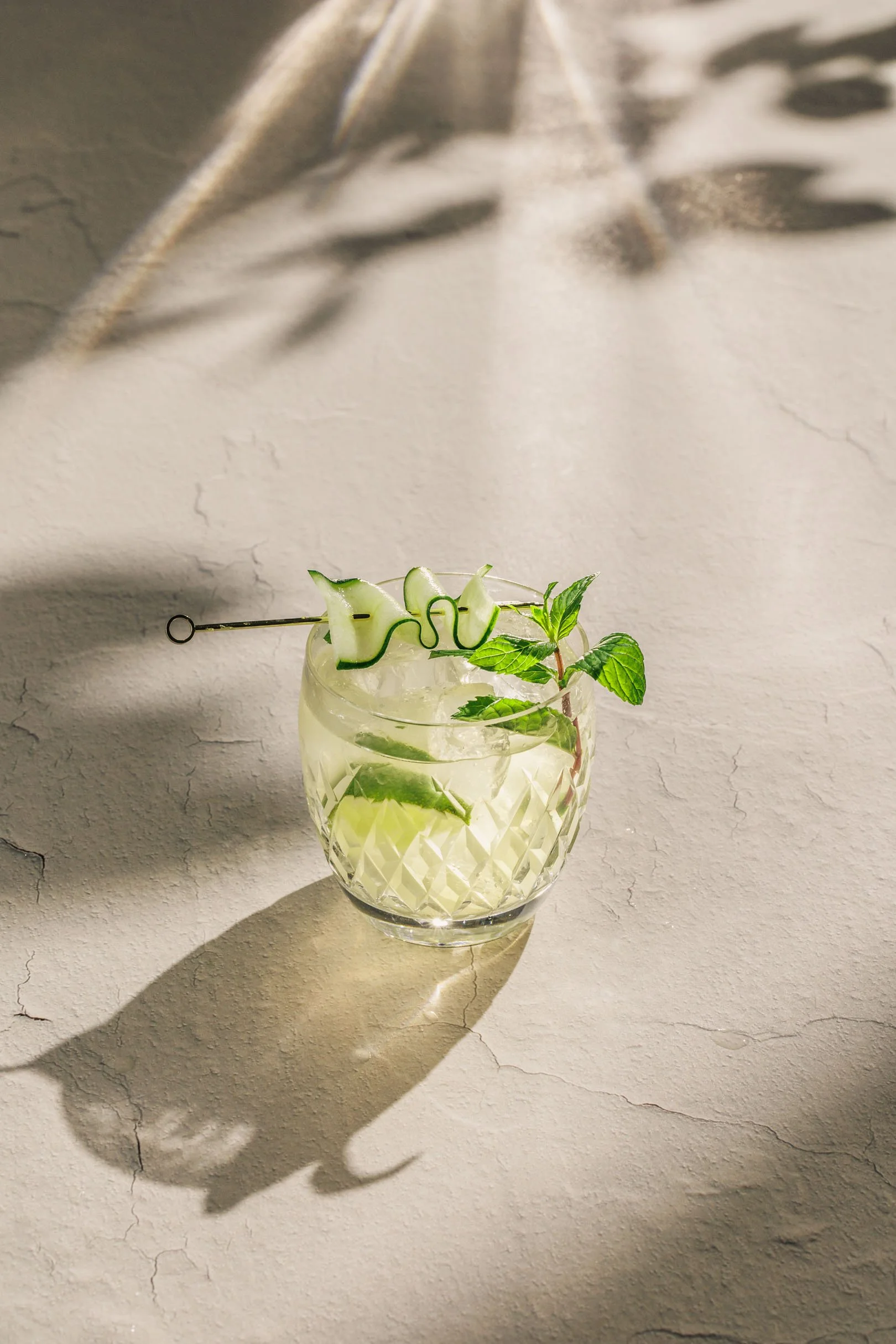 A glass of cocktail, mint julep with cucumber and mint garnishes on a textured surface, shadow cast by leaves