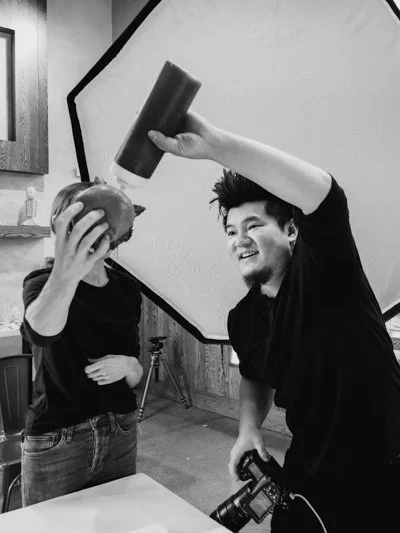 Two people are in a photography studio; one is holding an apple in front of their face, and the other is pouring a drink or condiment from a bottle over the apple. A camera is visible in the hand of the person pouring, and a large studio light is in the background.