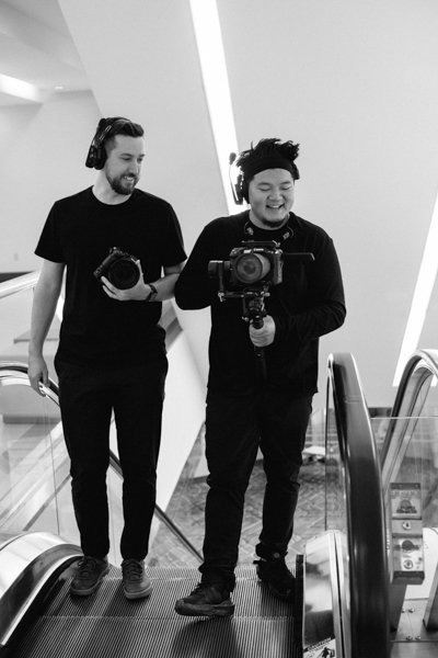 Two young men, both wearing headphones and black clothing, are on an escalator taking a selfie with a professional camera, smiling and looking at the camera.