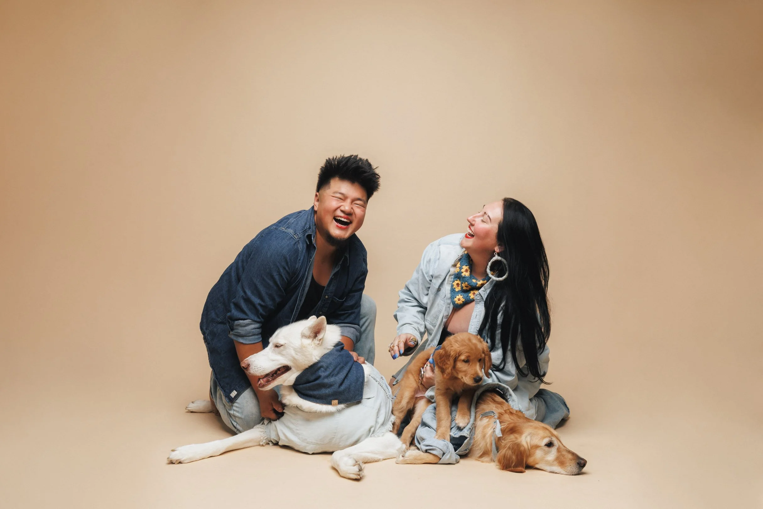 A happy couple sitting on the floor with their three dogs against a beige background. They are laughing and wearing casual denim jackets.