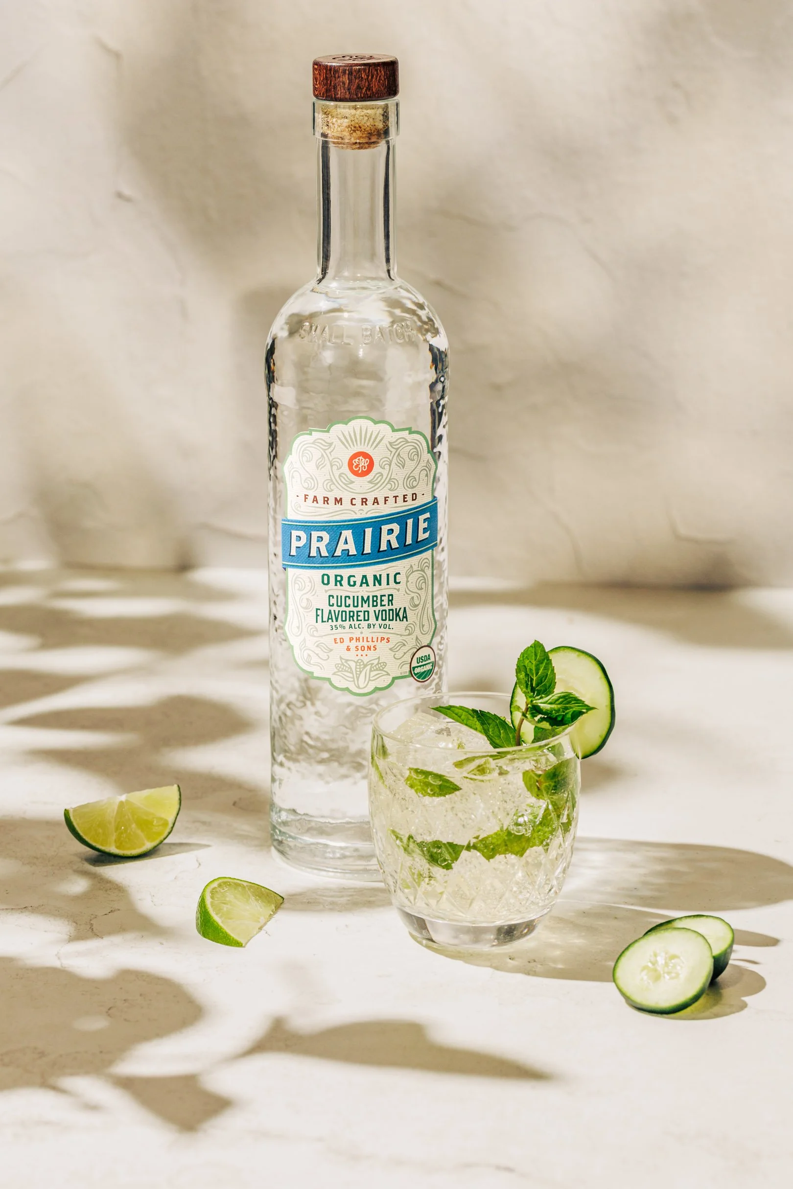 Bottle of Prairie organic cucumber flavored vodka with a glass of cucumber and mint-infused cocktail garnished with cucumber slices and mint leaves, along with lime wedges on a white surface.