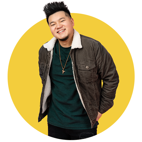 Young man with dark hair styled upward, wearing a black jacket with a white shearling collar over a dark green shirt, smiling and standing in front of a yellow background.
