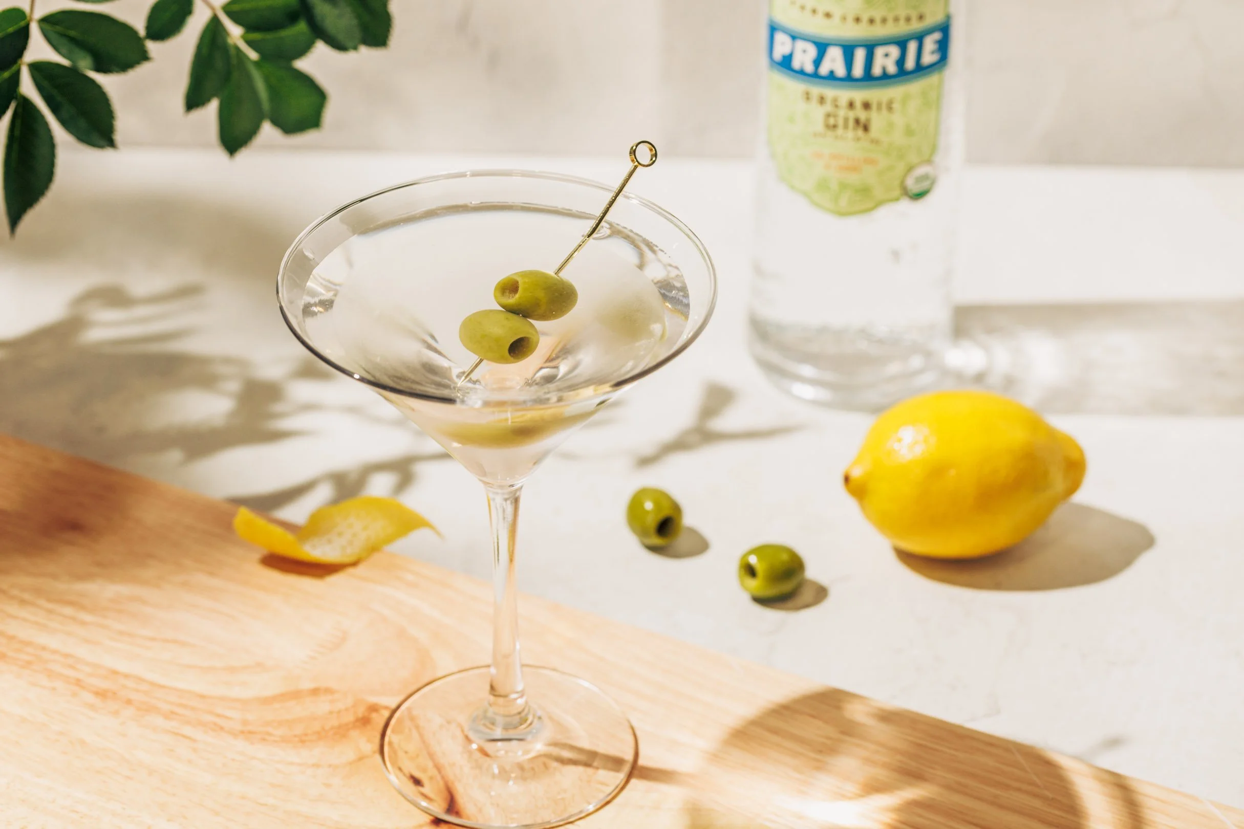 A martini glass with green olives, a lemon peel, and a cocktail stir stick on a wooden surface. In the background, there is a bottle of Prairie organic gin, a lemon, and some loose green olives on a white surface.