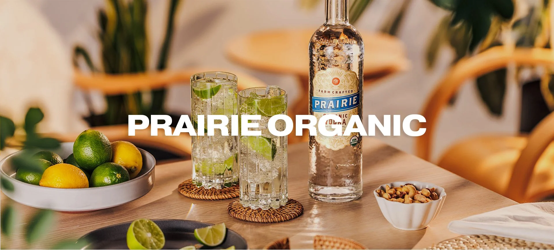 A bottle of Prairie Organic Spirit next to two glasses of lime and ice, surrounded by lemons, limes, pistachios, and lime wedges on a wooden table with a blurred background.