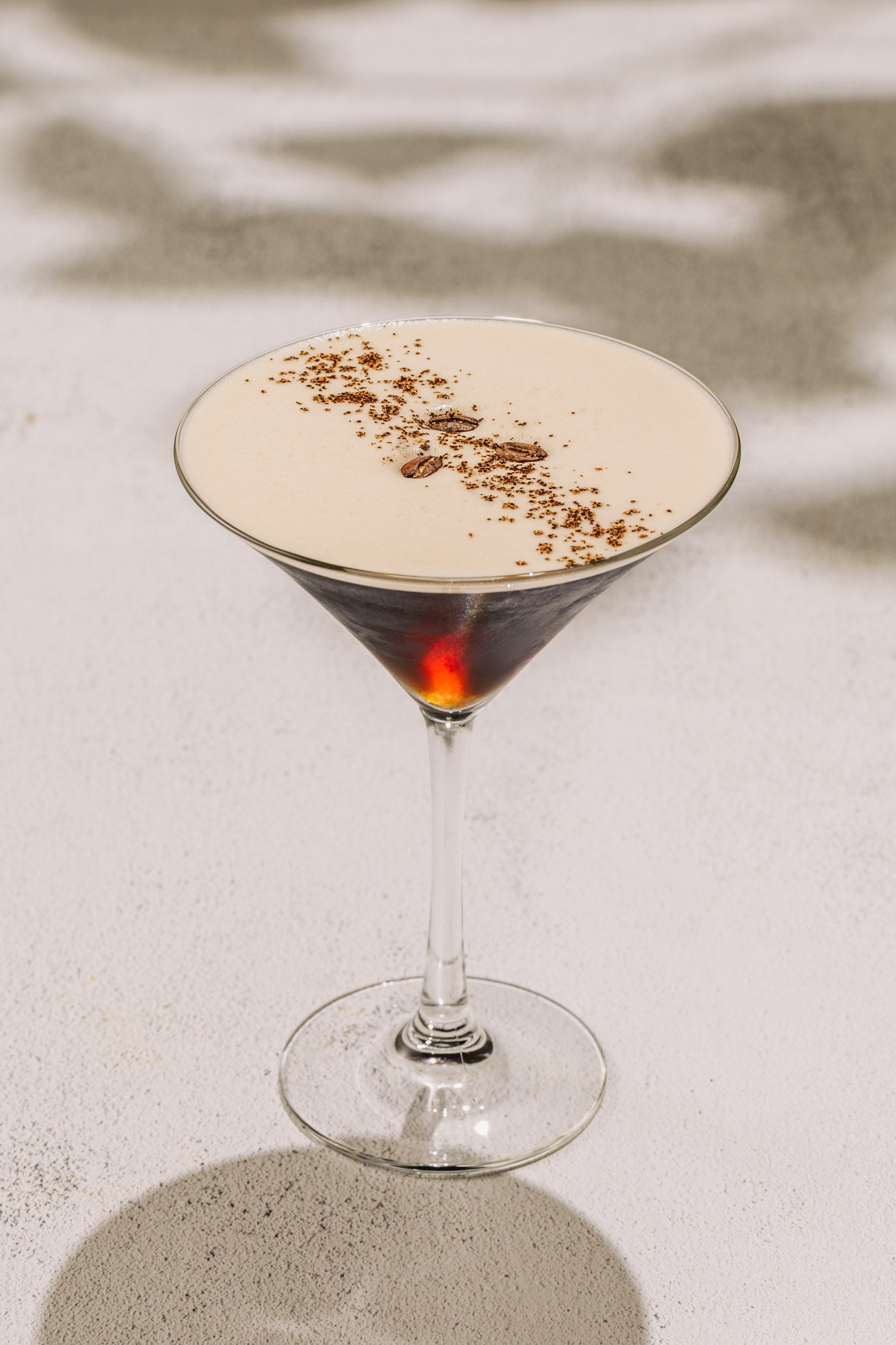 An espresso martini cocktail in a martini glass with a dark liquid layer at the bottom and a creamy white froth on top, garnished with coffee beans and cocoa powder.