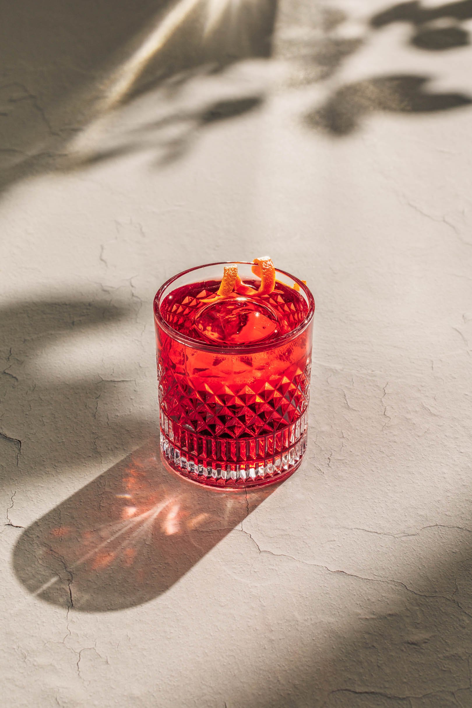 A red negroni cocktail in a textured glass with lemon peel garnish, casting a shadow on a light-colored textured surface with cracks.