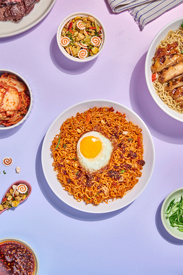A plate of spicy ramen noodles topped with a fried egg surrounded by various Asian dishes on a light purple background.