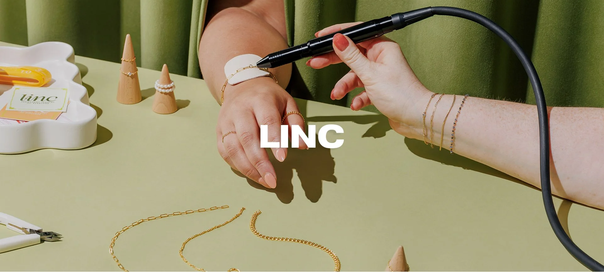 A person with fair skin is using a black handheld tool to embed a gold chain bracelet onto their forearm. The person is wearing multiple gold and beaded bracelets on their wrist and has a few gold rings on their fingers. On the table, there are jewelry pieces, including chains and rings, along with jewelry display cones and a white dish with tags and small tools. The background features a green curtain or wall.