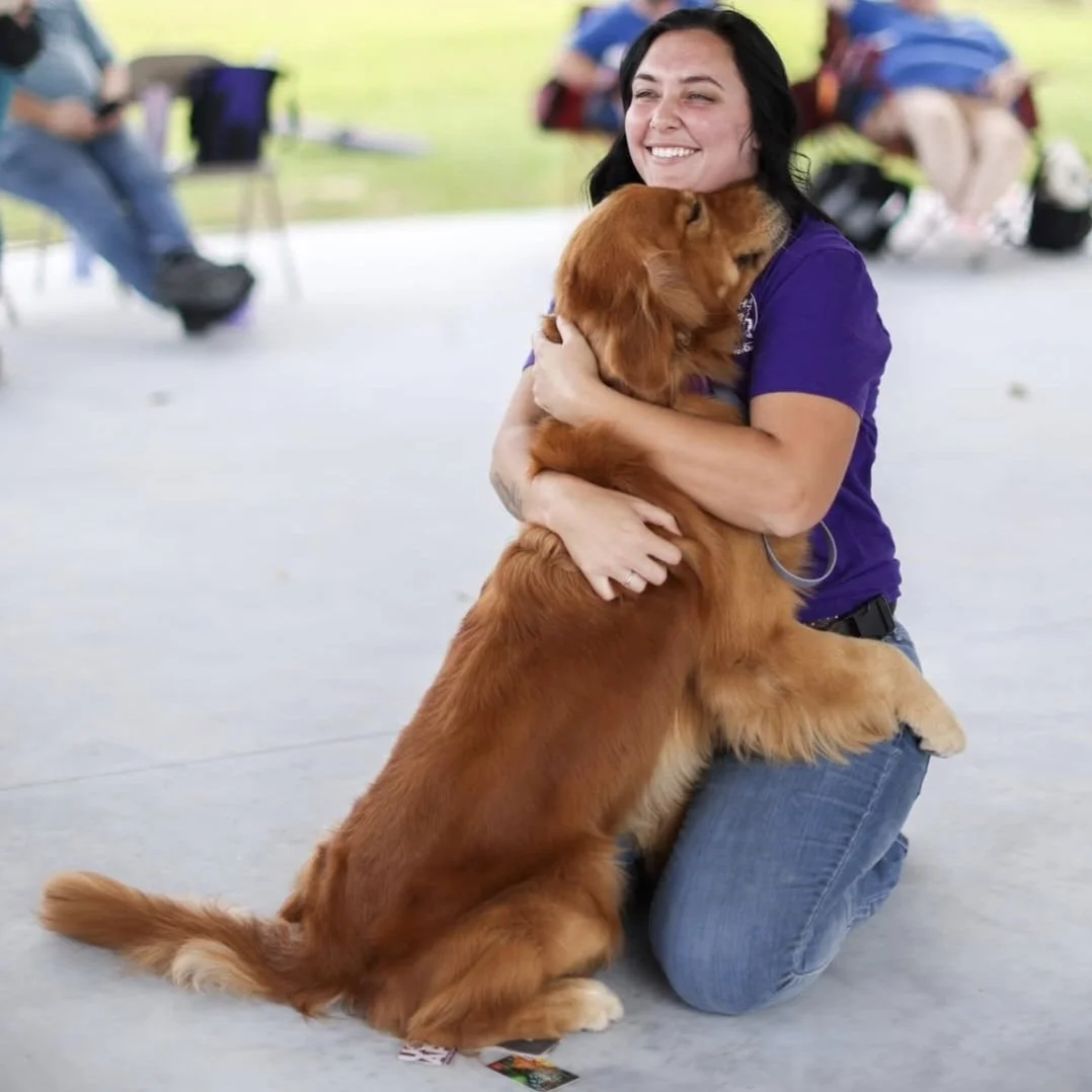 A woman kneeling on the ground, hugging a large golden retriever. The woman is smiling, and the dog is standing on its hind legs, leaning into her embrace. The background shows other people sitting and standing, outdoors in a park or similar setting.