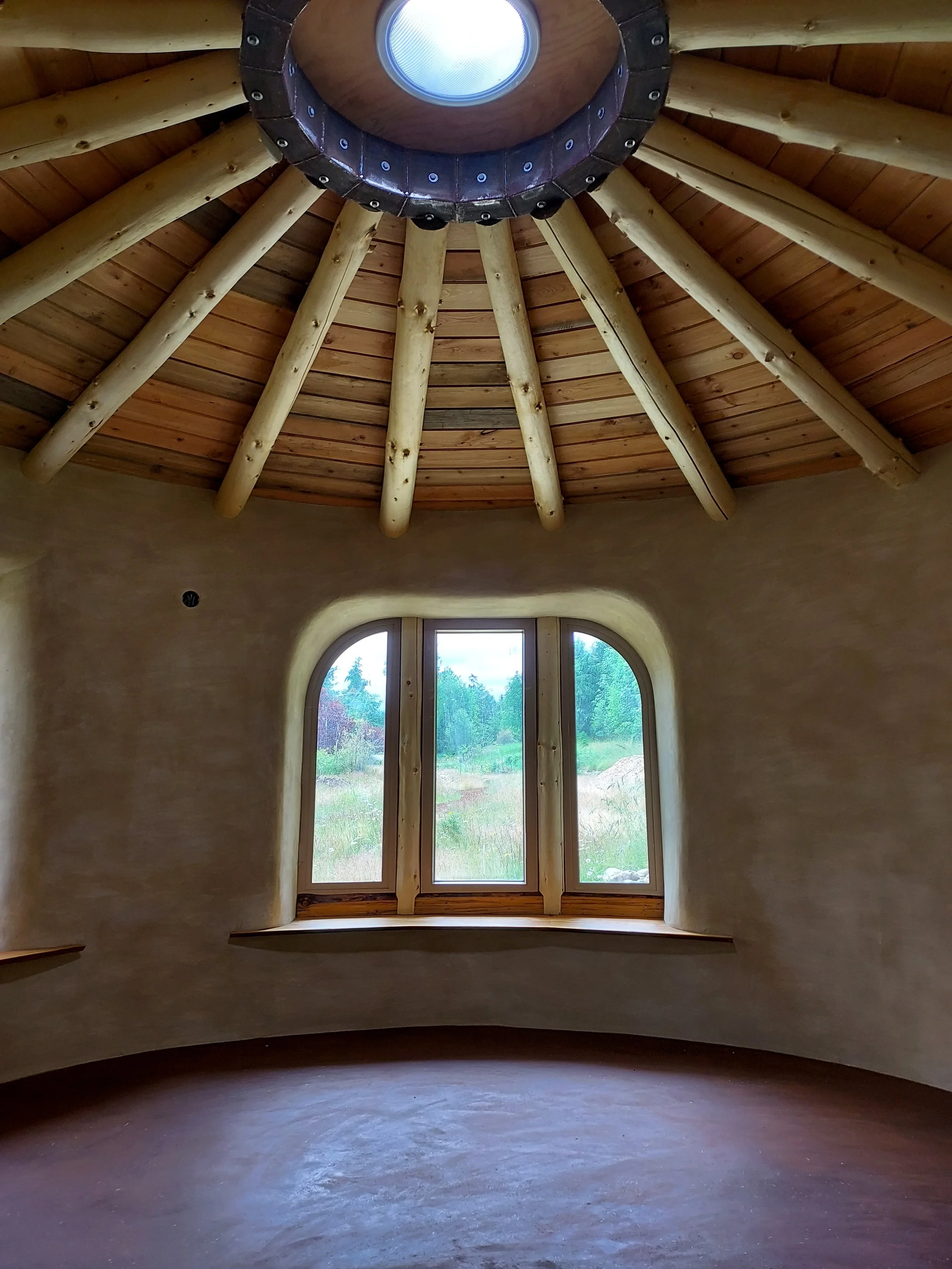 Interior view of a room in a natural home with smooth living clay walls and a high wooden ceiling and large arched window.