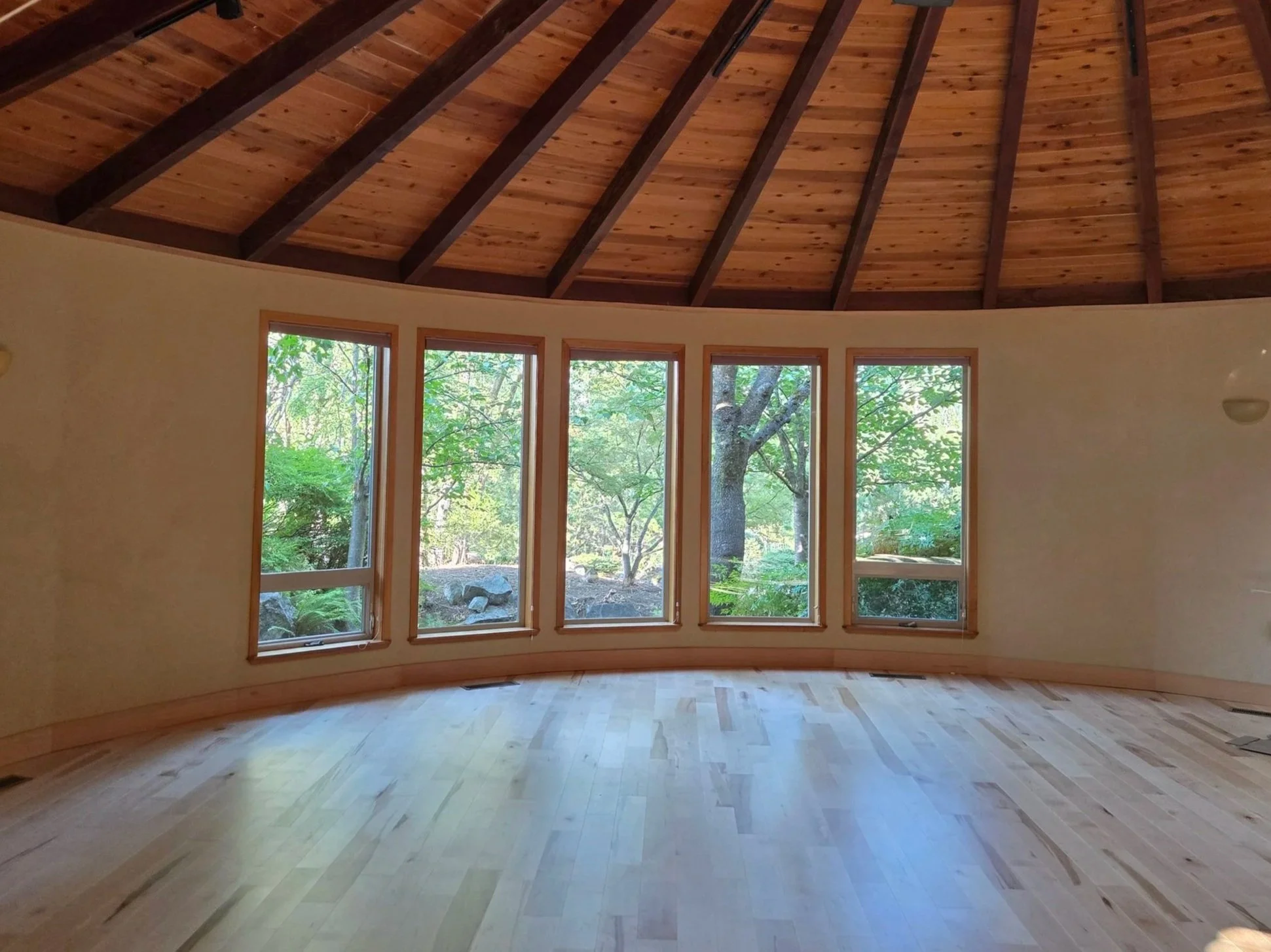Inside view of a circular room with large windows showing a green wooded outdoor scene, light wood floors, and a wood-paneled arched ceiling.