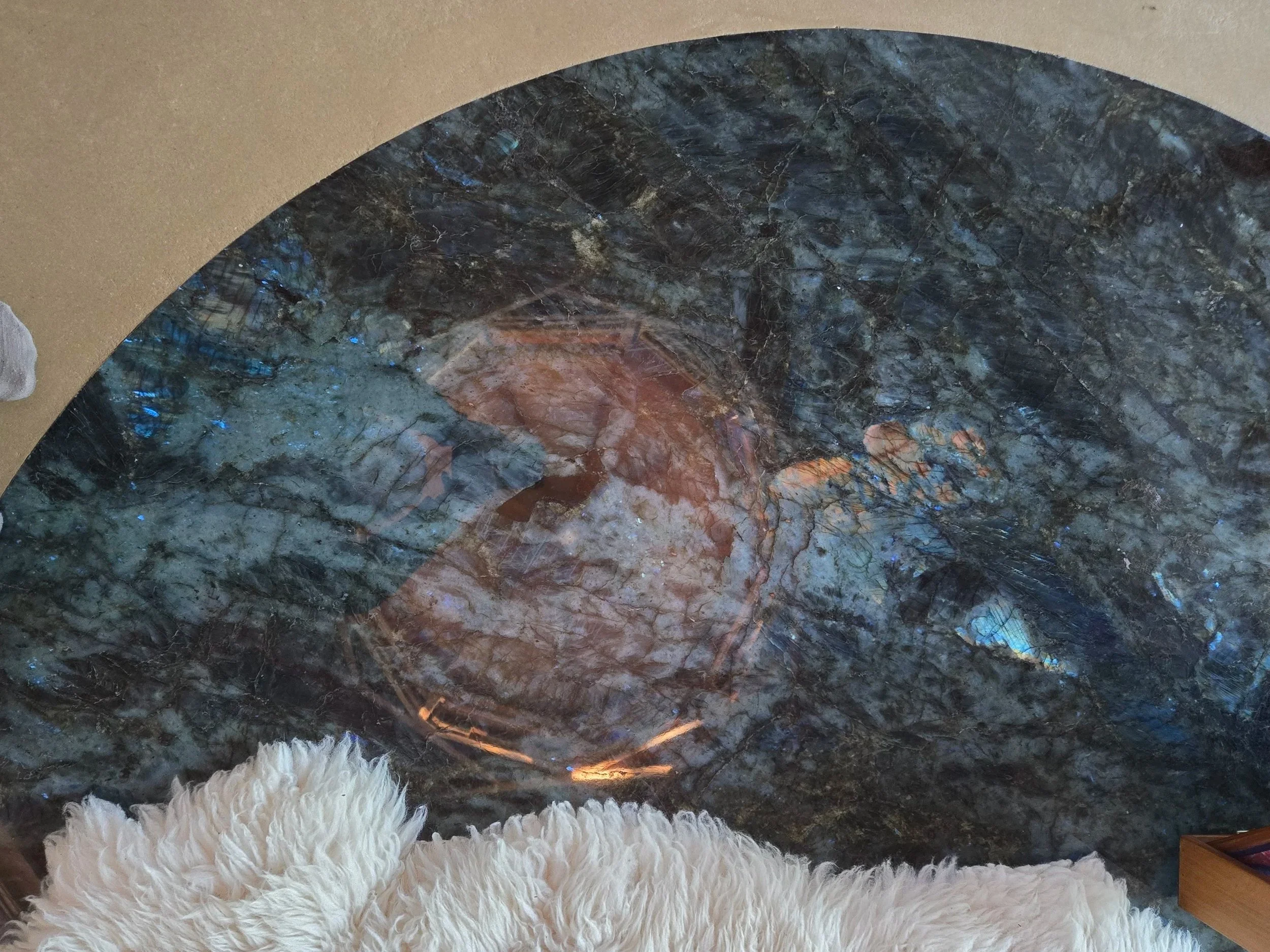 Close-up of a round table with a dark, multi-colored, polished stone surface and a white fluffy rug in front of it.