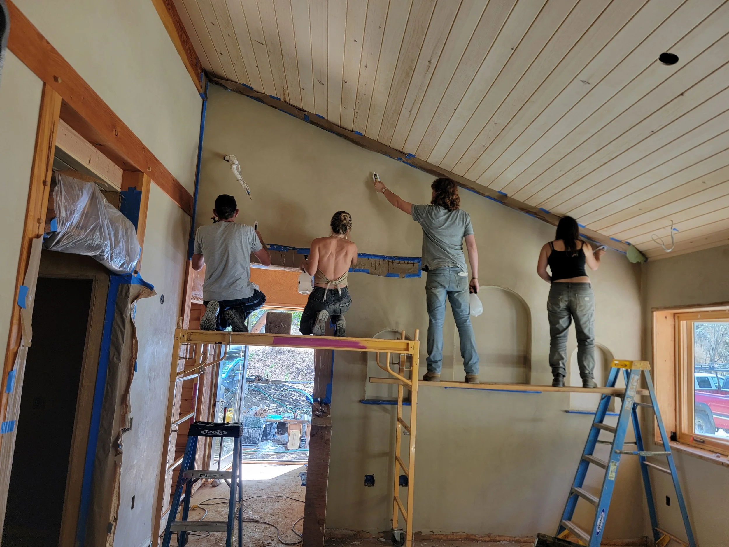 Four people are working on painting or finishing a wall in a room under renovation. They are standing on scaffolding and a ladder, with one person using a paint roller, two others holding paint brushes, and one woman looking at the wall. The room has