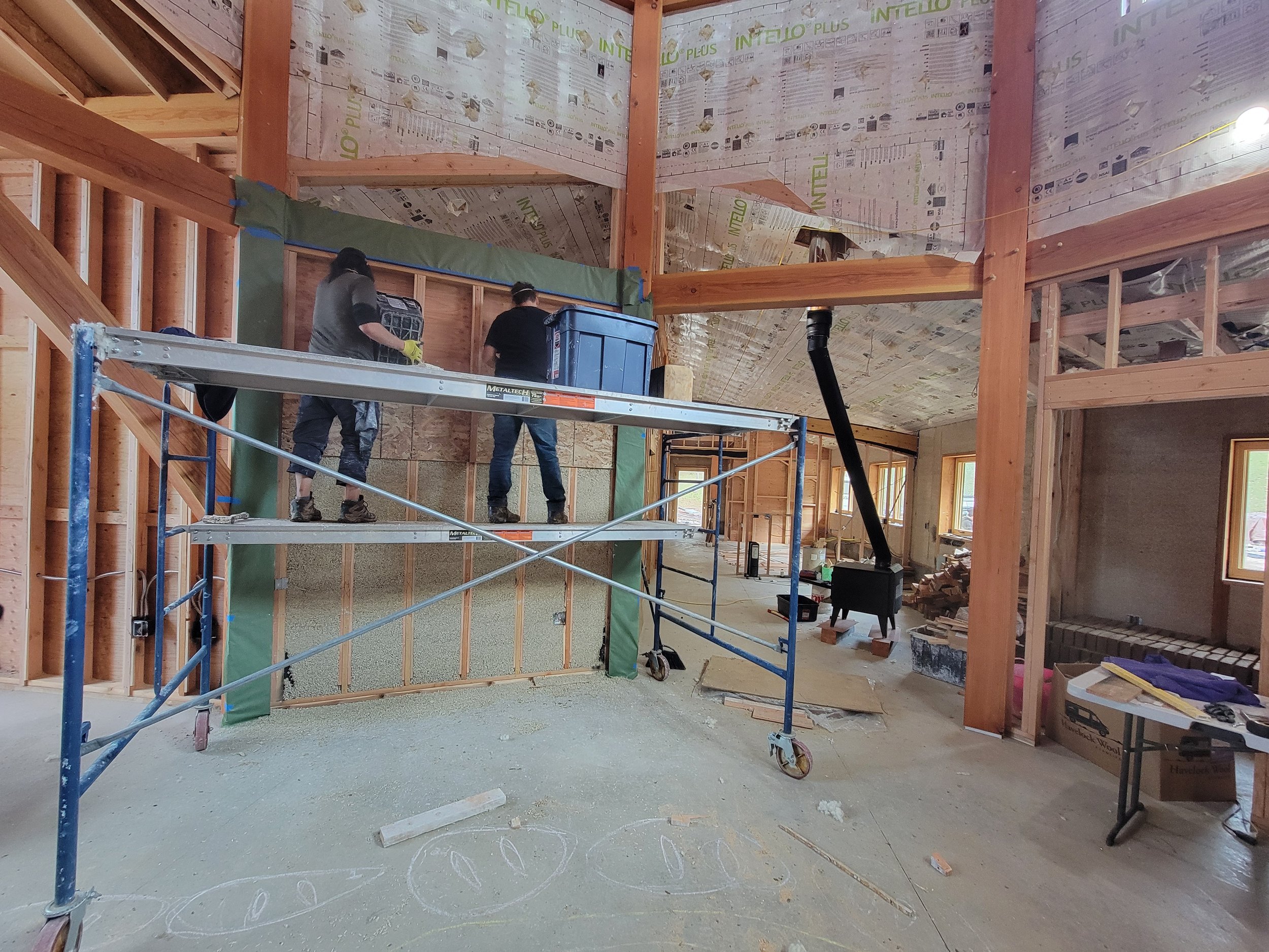 Interior view of a house under construction showing workers on scaffolding installing insulation on a wall. The space features exposed wooden beams and a high ceiling.