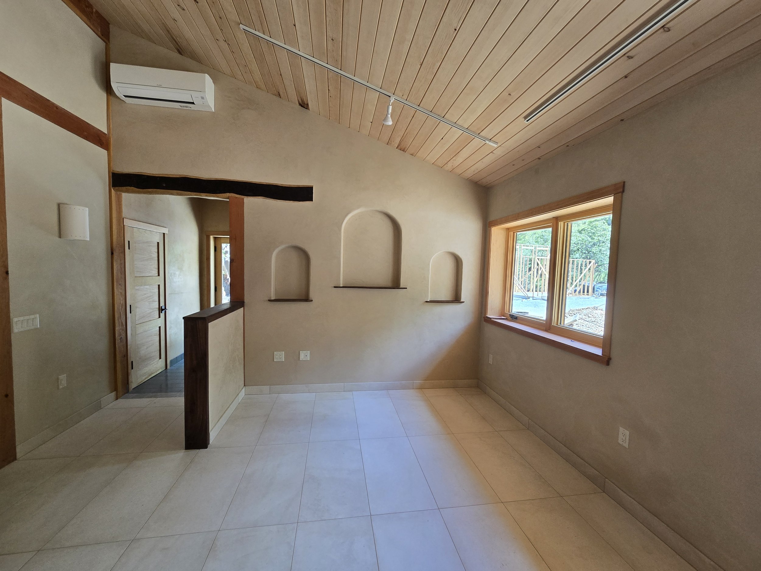 Empty room with beige walls, wooden ceiling, tiled floor, and a large window showing an outdoor construction site. Niche shelves are built into the wall.