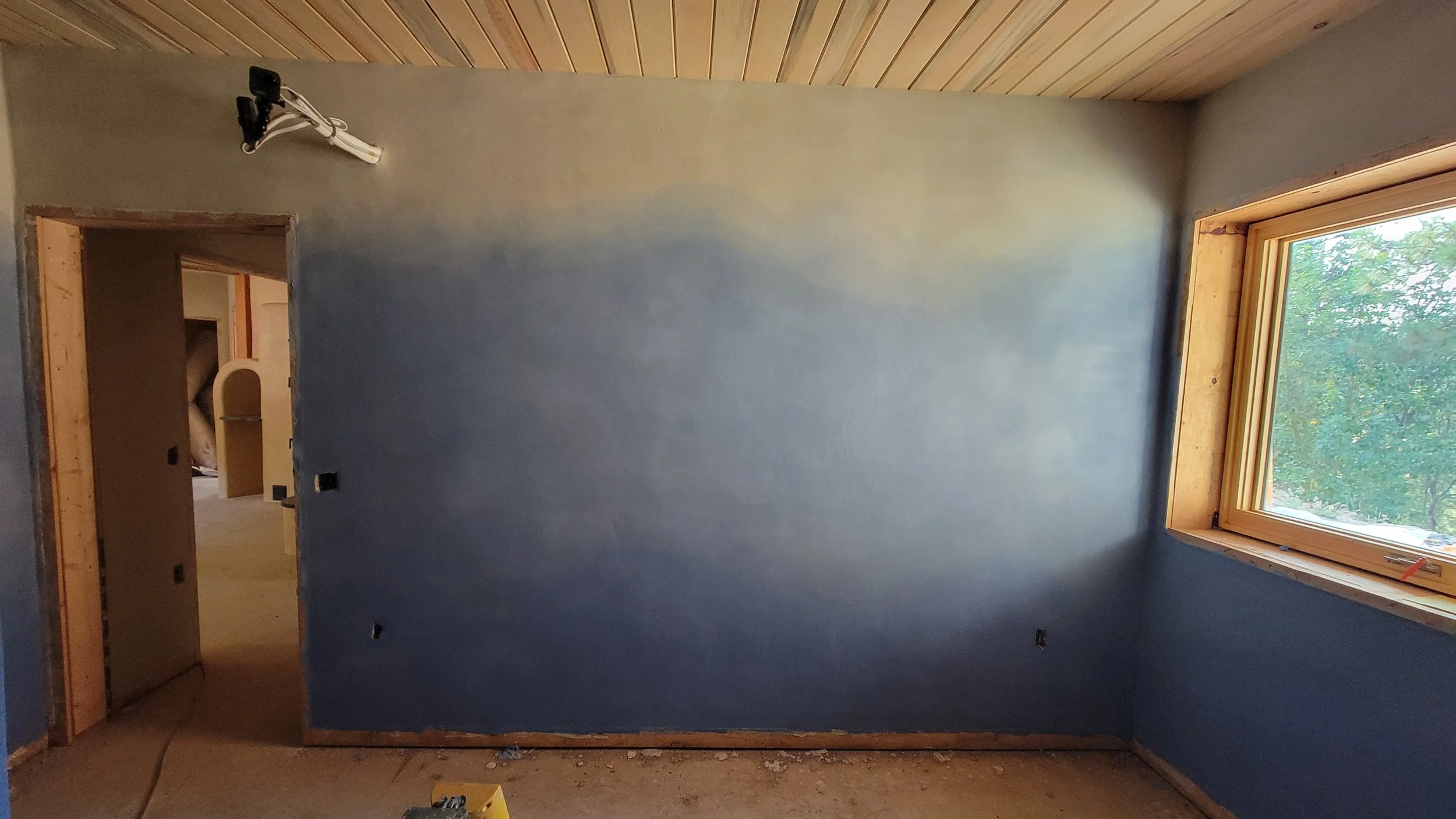Room under construction with a blue wall, window showing trees outside, and an open doorway leading to another room.