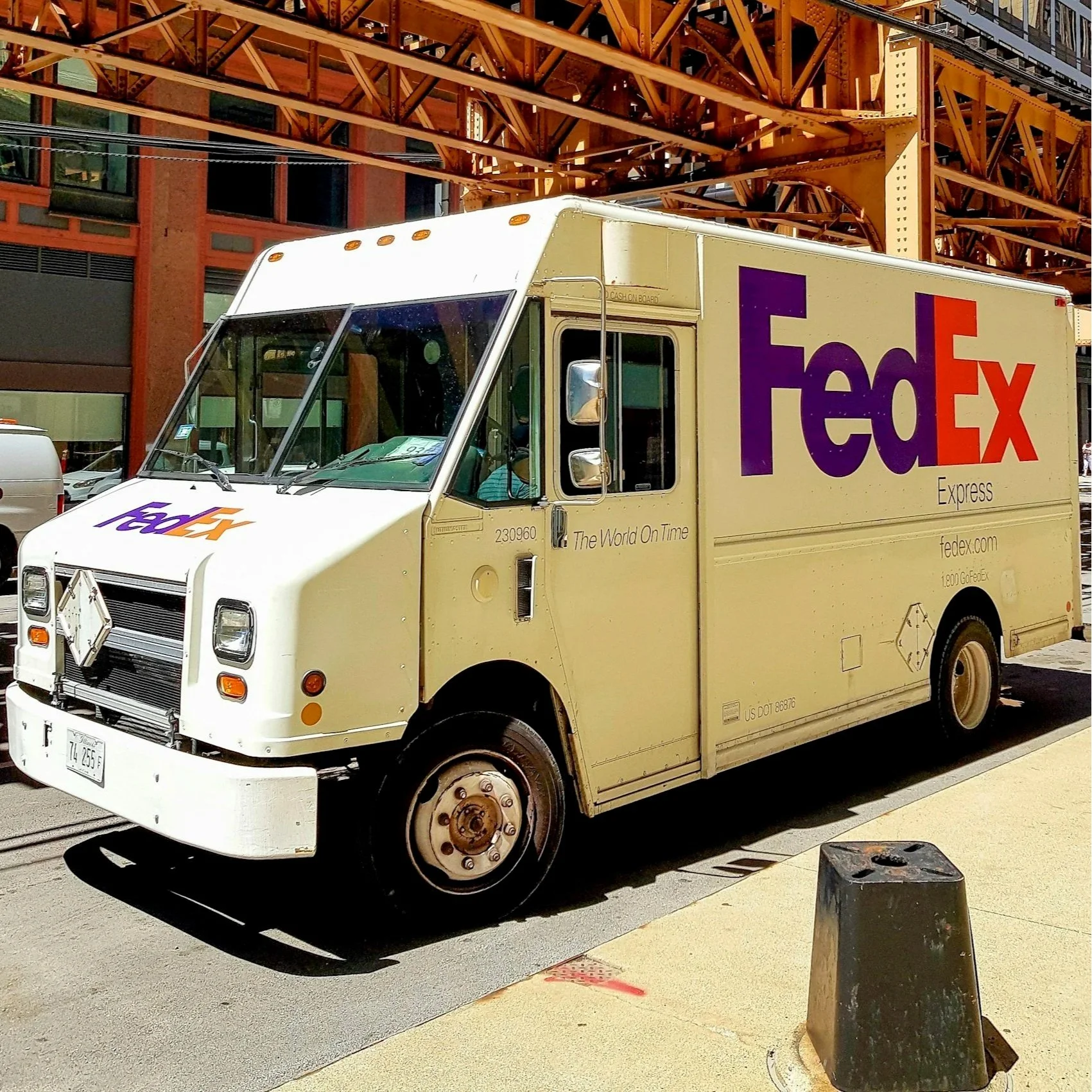 FedEx delivery truck parked under an orange steel bridge near a building with large windows