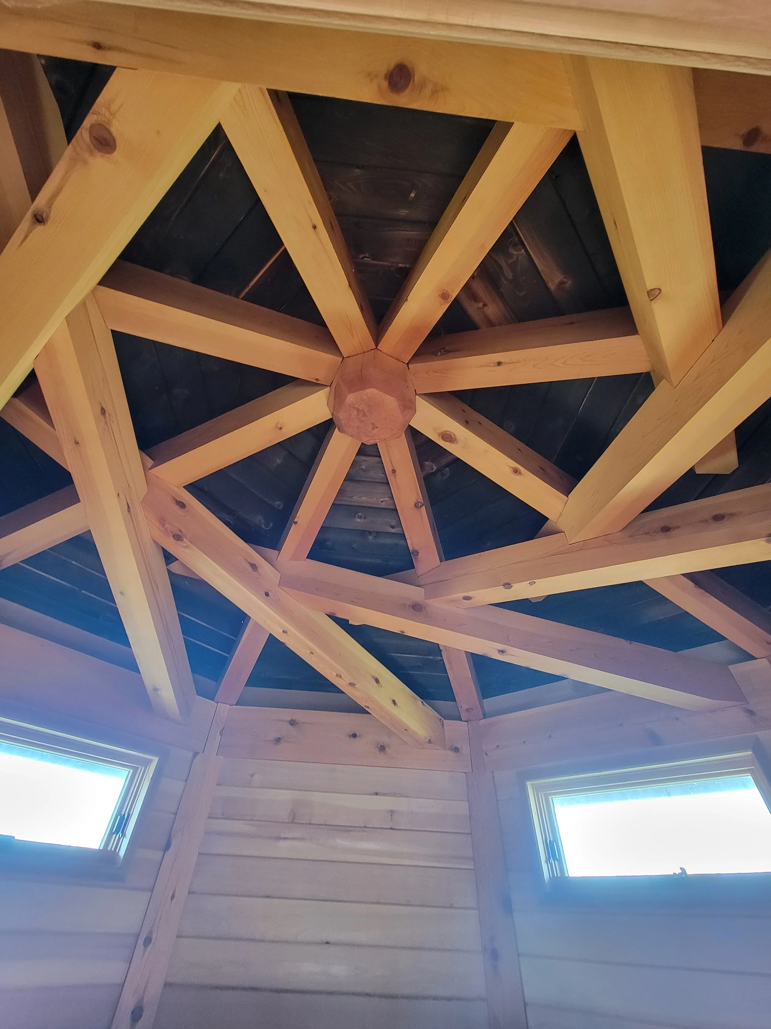 Interior view of a wooden ceiling with a geometric pattern, featuring light wood beams and black wood panels, and two small rectangular windows.