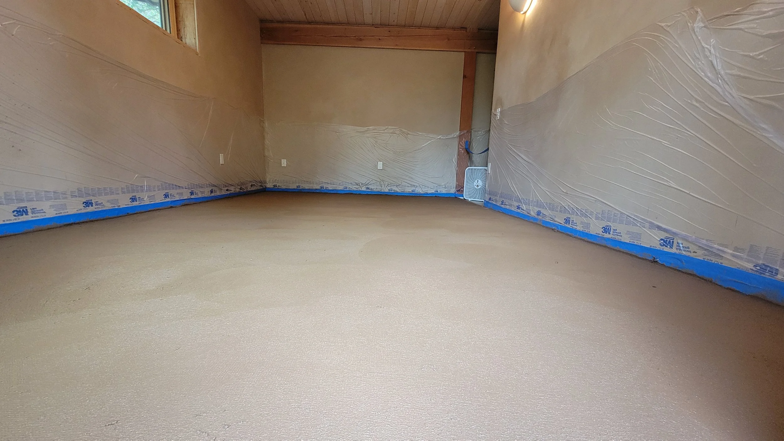 Empty room with beige walls, plastic sheeting on the walls, and a beige textured carpeted floor. There is a small white fan plugged into an outlet in the corner and a window on the left side.