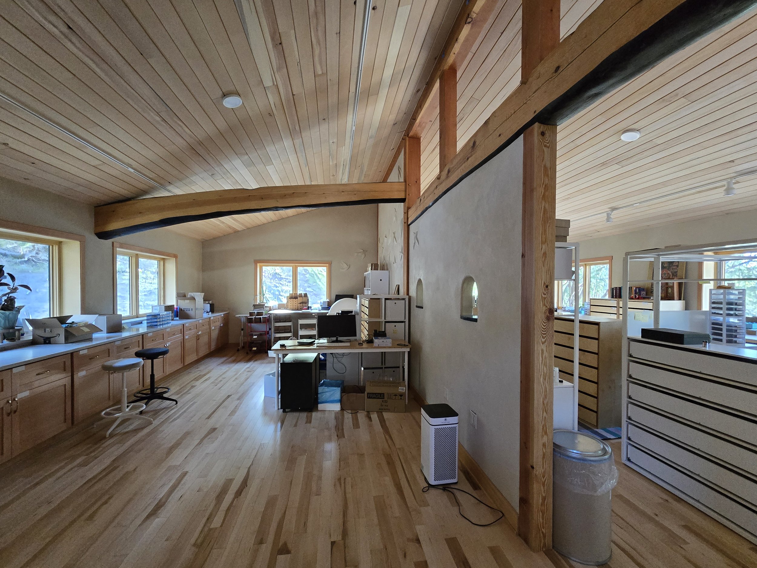 Interior of a room with wooden floors and ceiling, featuring desks, chairs, and shelves with windows revealing a natural landscape outside.
