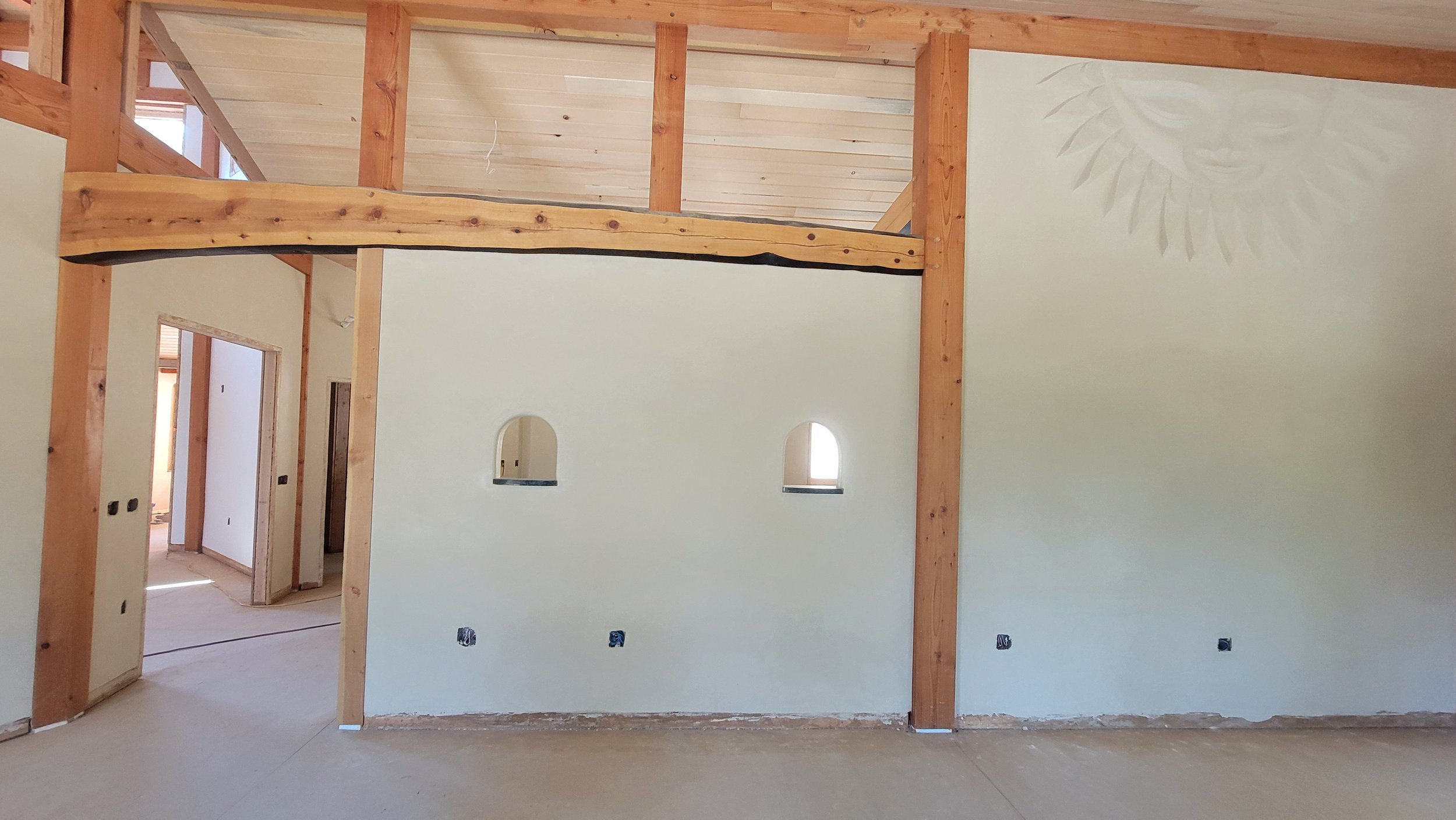 Interior of a house under construction with wooden beams, white walls, small arched windows, and a partially visible doorway.
