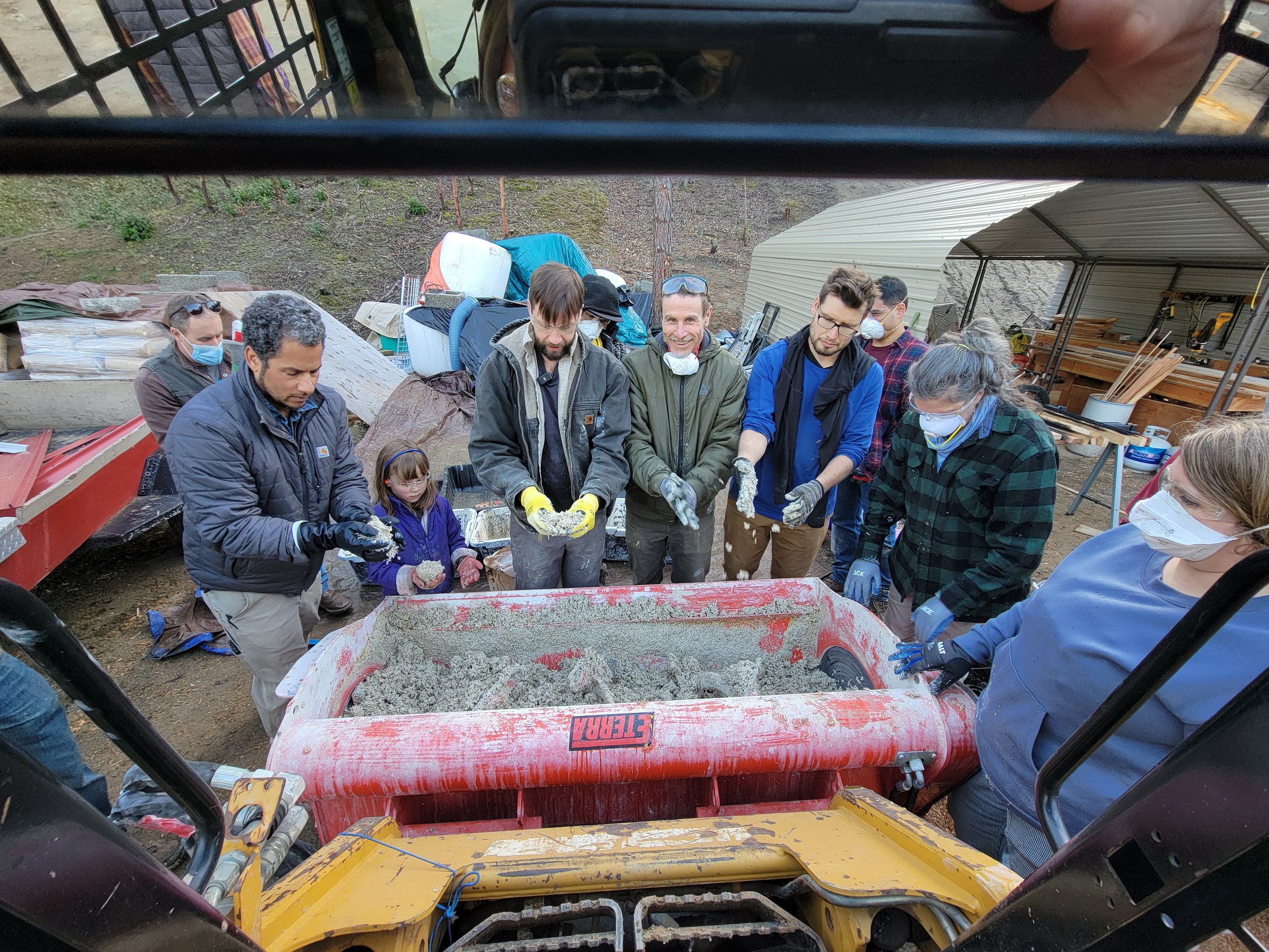 Group of people at a construction site working with cement or concrete, surrounded by tools and equipment, all wearing masks, gloves, and outdoor clothing.