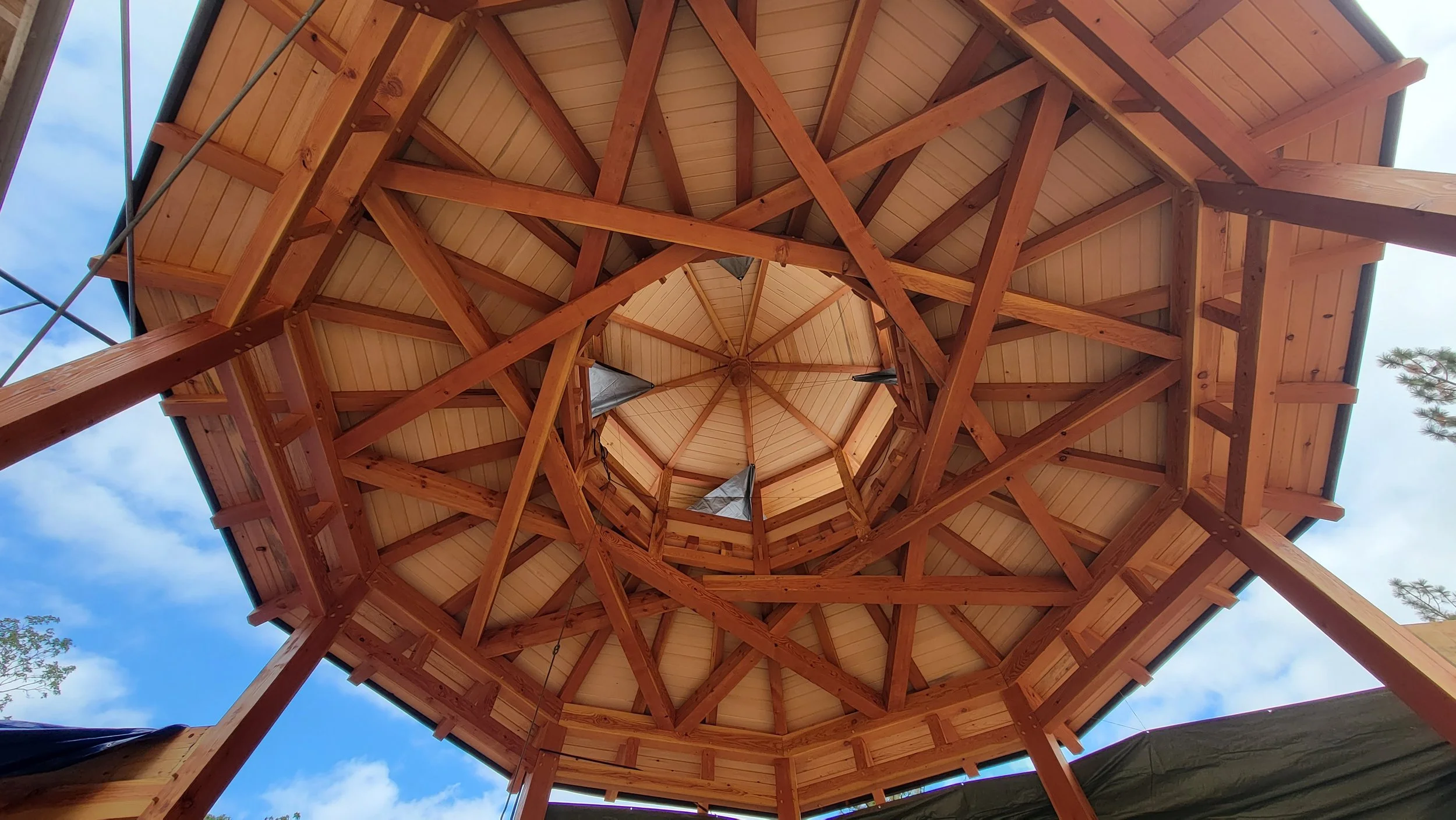 Looking up inside a wooden gazebo with a hexagonal structure, showing wooden beams and panels forming a spiral pattern toward the center, with a partly cloudy blue sky visible through openings.