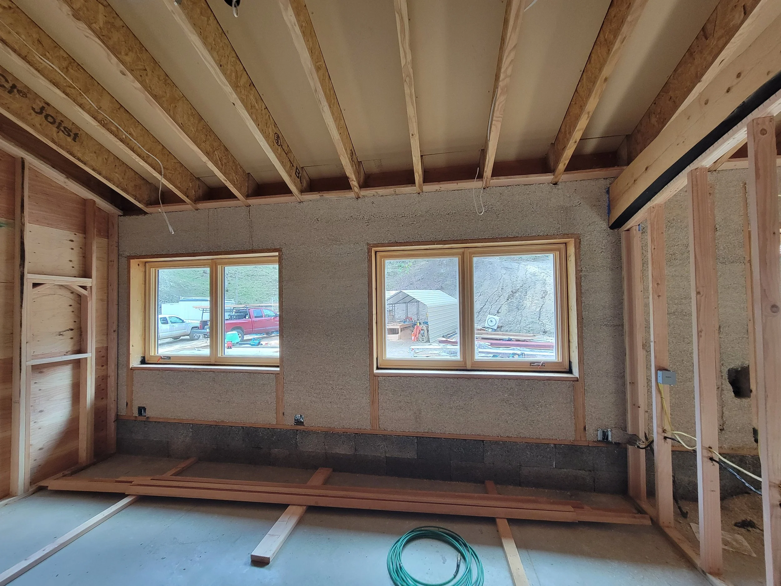Interior of a house under construction with framed walls, an unfinished ceiling with exposed wooden joists, two windows, and construction materials on the floor.