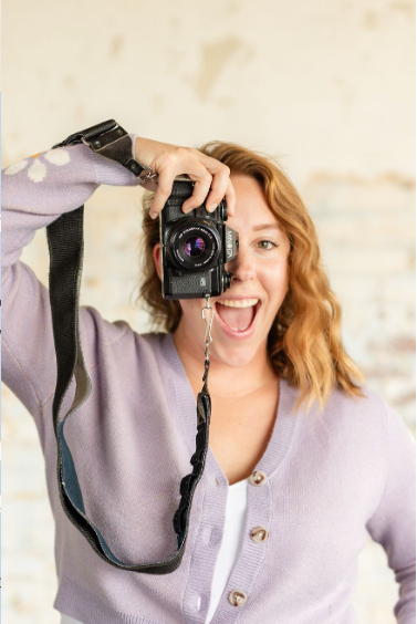 A woman with red hair smiling and holding a camera up to her face, taking a picture.