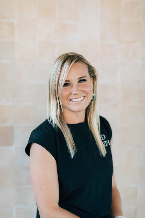 A smiling woman with shoulder-length blonde hair, wearing a black t-shirt, standing against a beige tiled wall.