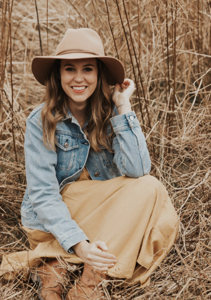 A woman in a denim jacket, tan skirt, and brown boots sitting in a field of dry grass, wearing a beige wide-brimmed hat and smiling.