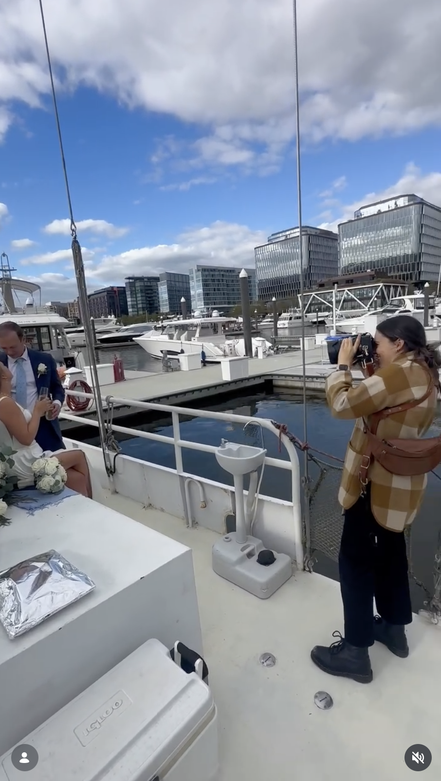 A woman taking a photograph of a couple during a wedding ceremony on a boat at a marina with yachts, modern office buildings, a blue sky with clouds in the background.