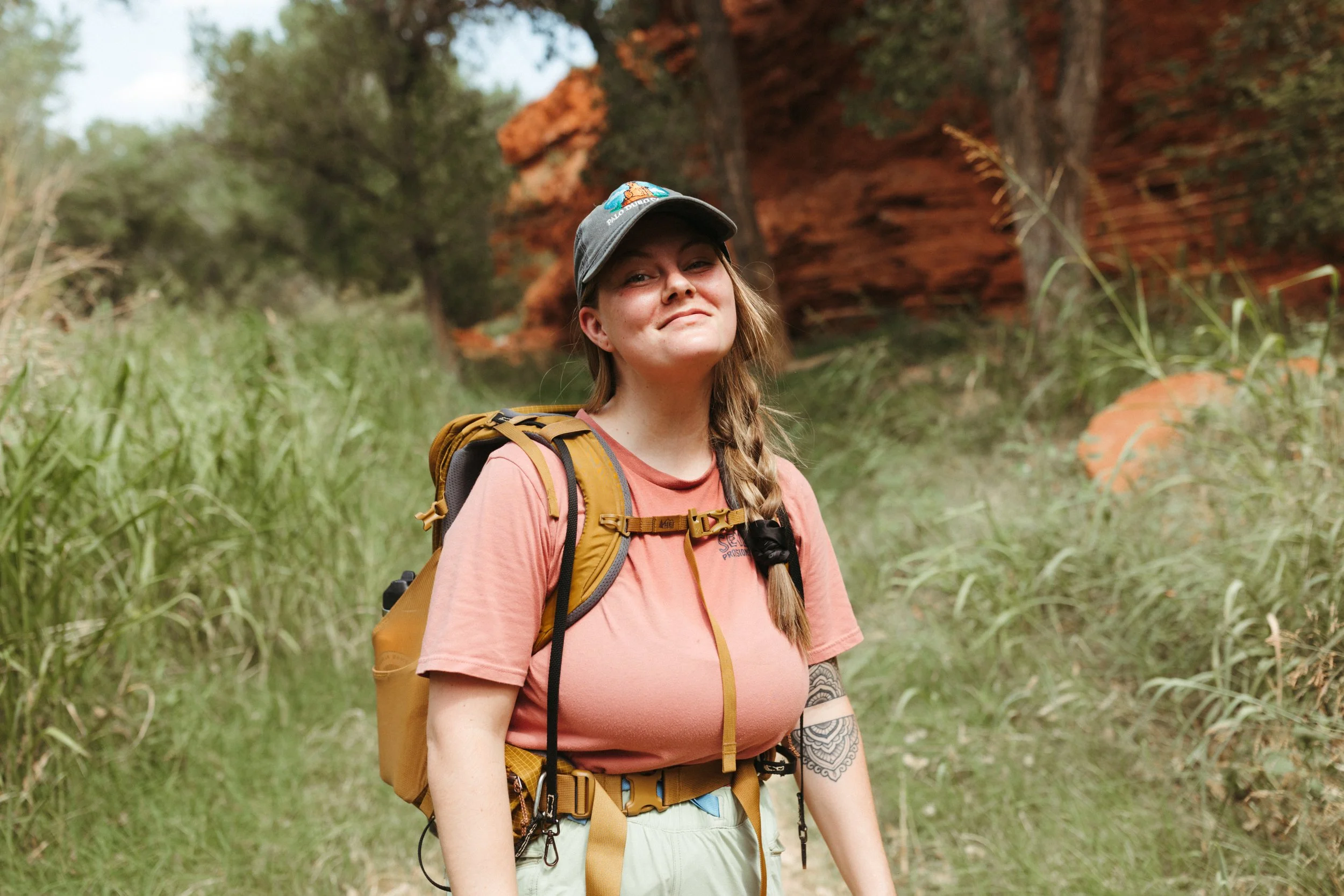 Young woman with long hair in a braid, wearing a gray cap, pink T-shirt, and carrying a yellow backpack, standing in a forest with red rock formations and green trees in the background.
