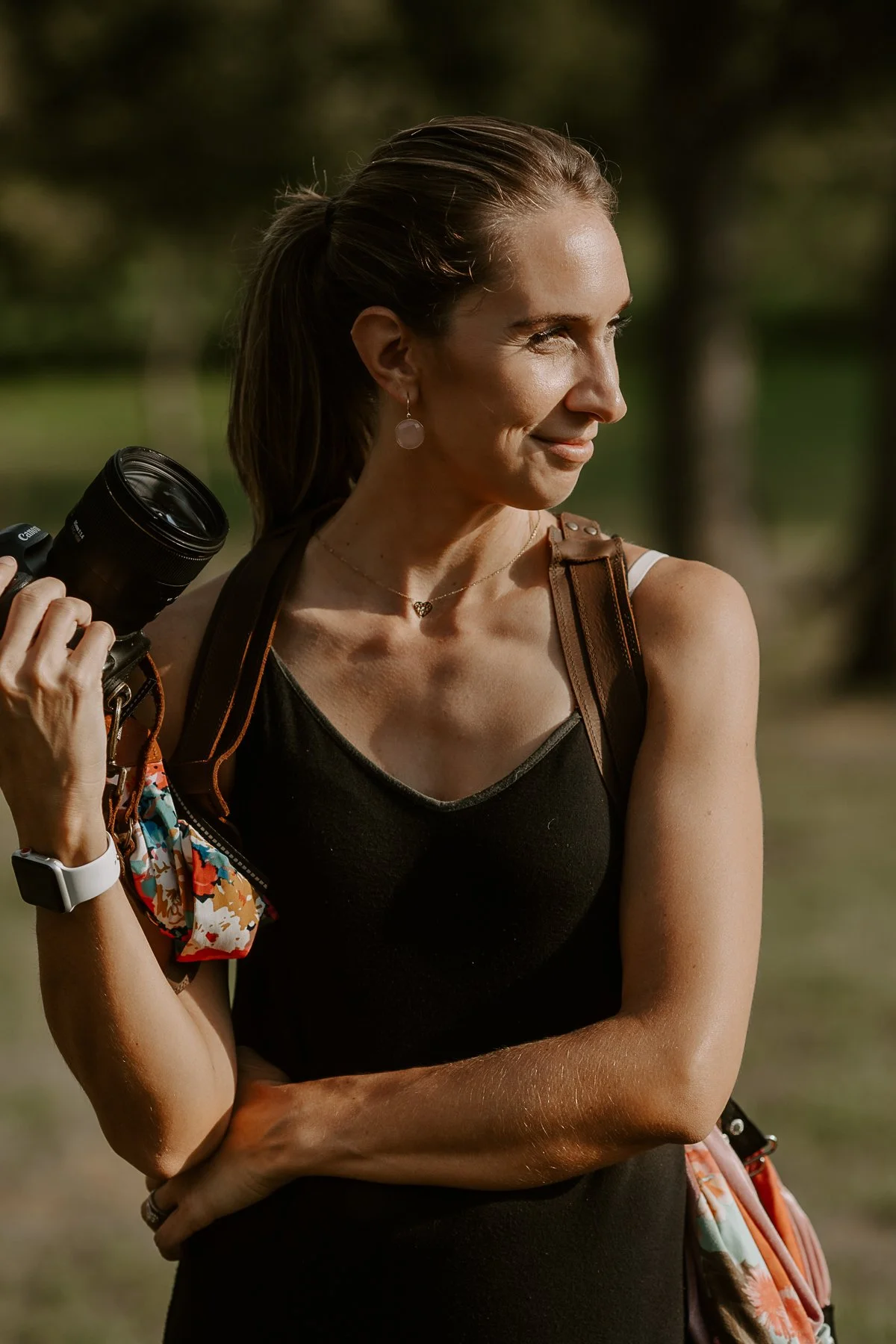 A woman outdoors with a camera slung over her shoulder, looking to the side with a slight smile, wearing a black top, earrings, a necklace, and a colorful bag.