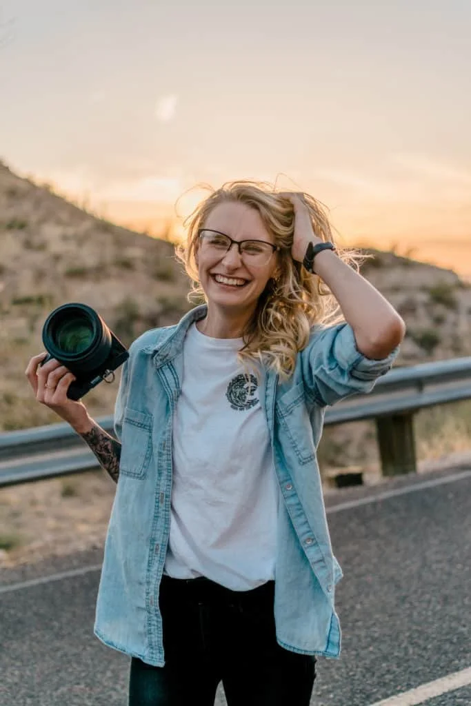 Smiling woman with glasses, holding a camera, standing on a road with a guardrail in a hilly outdoor setting at sunset.