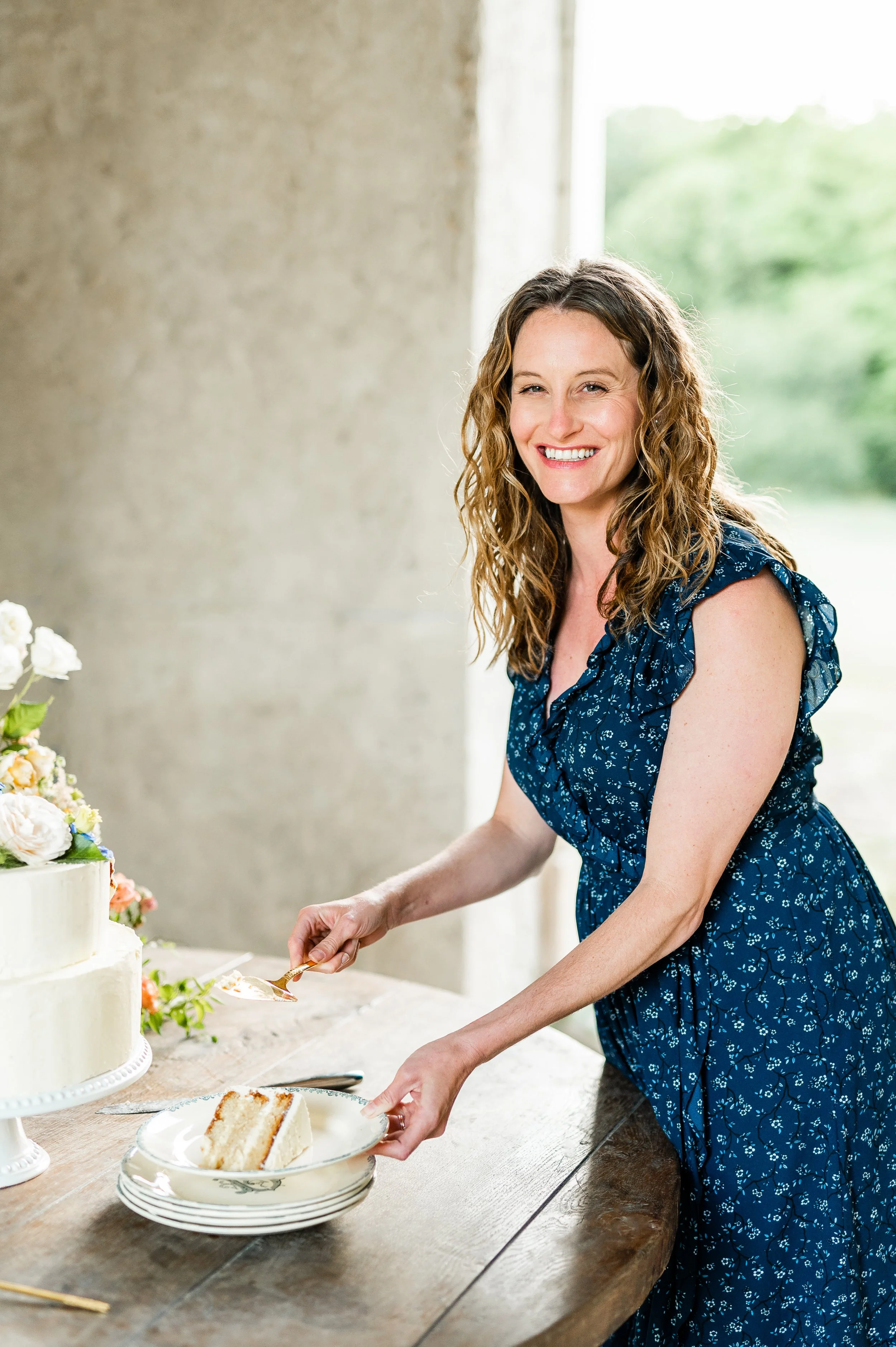 A woman in a blue floral dress smiling and cutting a slice of cake at a celebration or gathering, with a tiered cake and floral decorations visible.