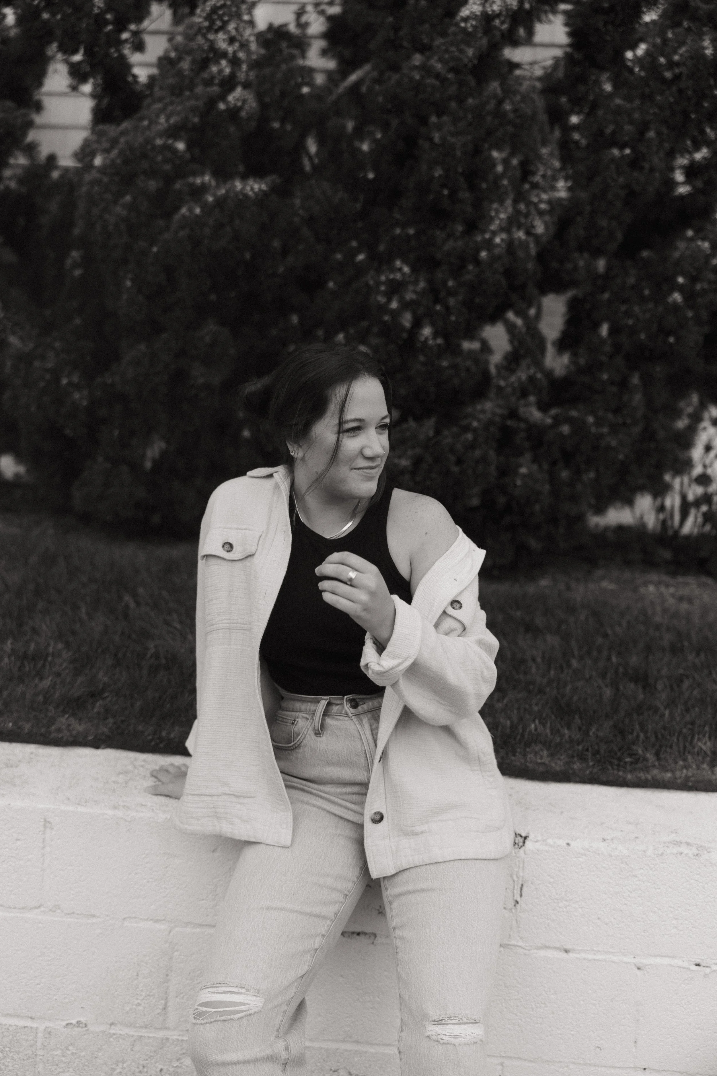 A young woman with dark hair styled in a bun, smiling slightly, looking to her right. She is wearing a black sleeveless top, a light-colored jacket draped over her shoulders, and ripped jeans. She is outdoors, sitting on a ledge with a background of large trees and a building.