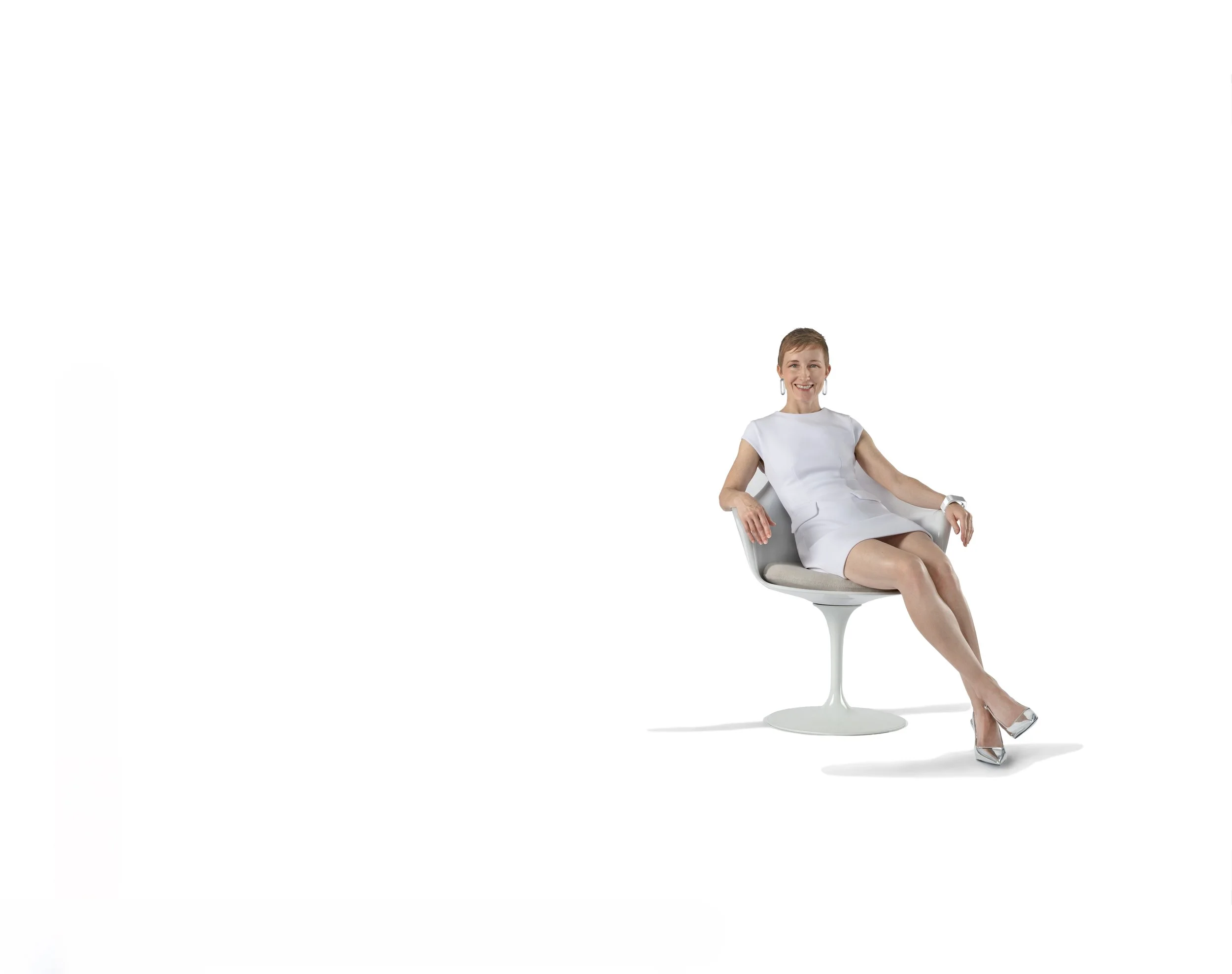 A woman sitting in a modern white chair, wearing a white dress and silver high heel shoes, smiling, with short brown hair and hoop earrings, against a plain white background.